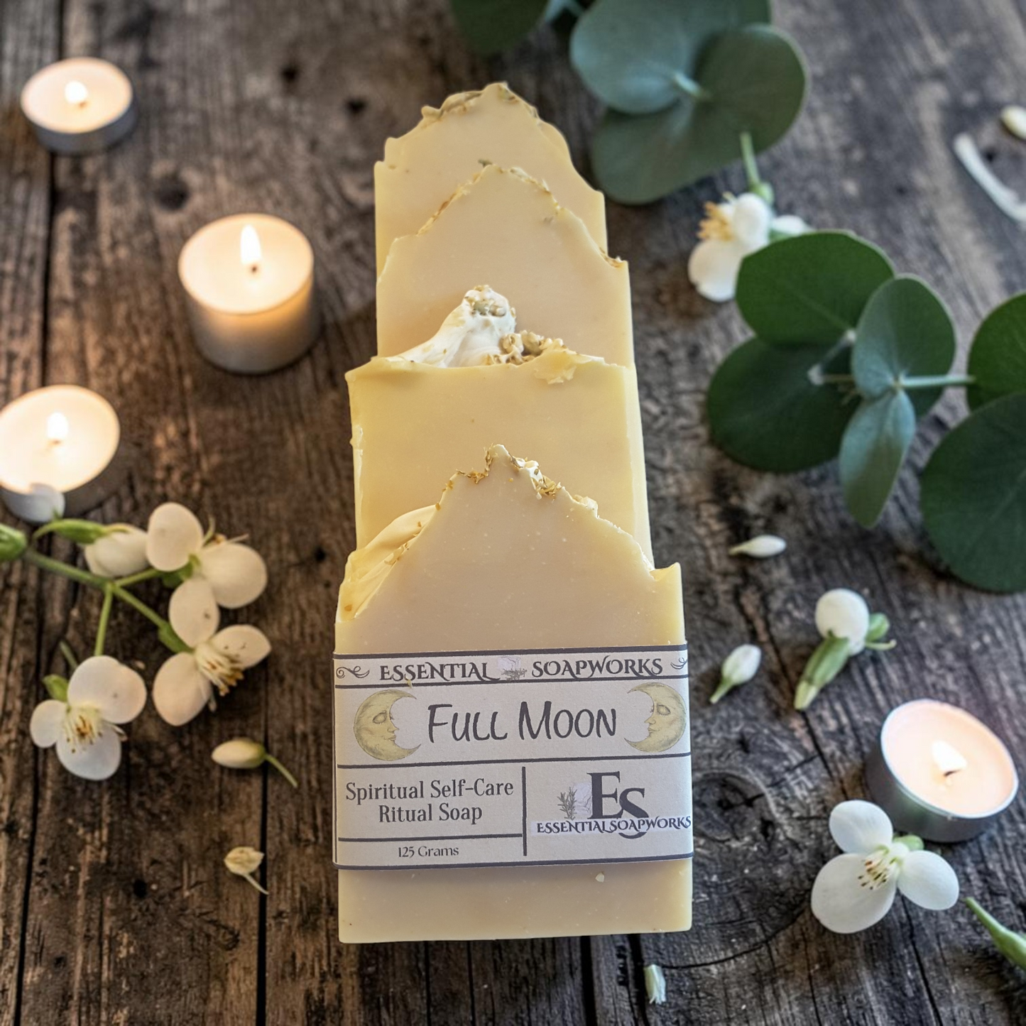 Four bars of Essential Soapworks Full Moon Ritual Soap arranged upright on a rustic wooden surface, surrounded by lit tea lights, white flowers, and green eucalyptus leaves.