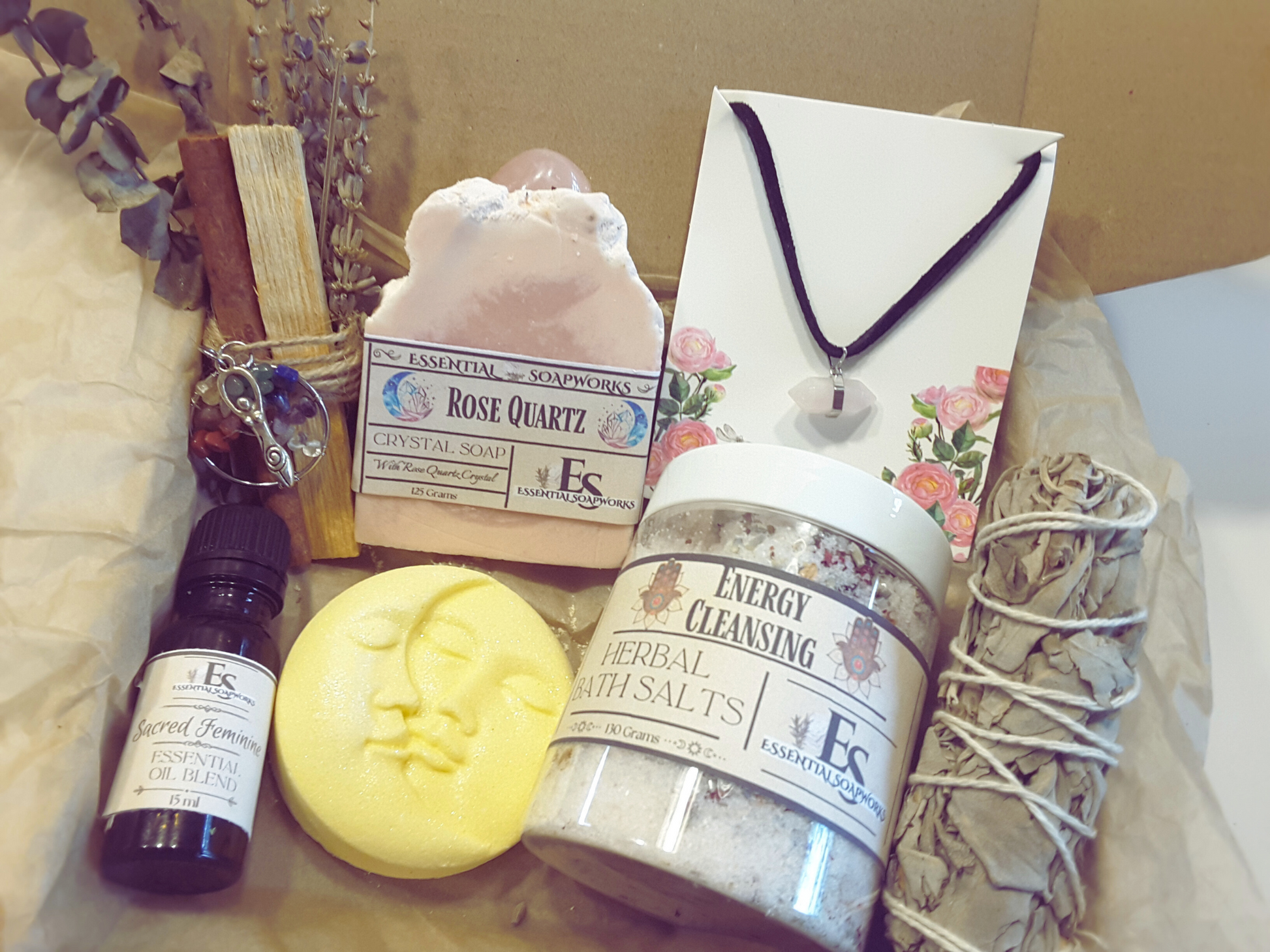 rose quartz mind, body and spirit self-care kit featuring a vegan soap topped with a rose quartz crystal, herbal bath salts, sacred feminine essential oil blend, rose quartz necklace, sun and moon bath bomb, white sage smudge stick and palo santo, cinnamon, lavender and eucalyptus smoke cleansing stick with a crystal tree of life charm and triple goddess charm