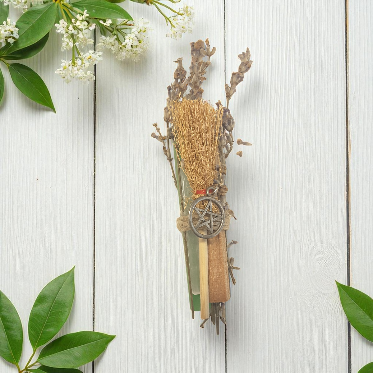 Decorative broom with pentacle charm, selenite, sandalwood and chime candle on a white wooden background with green leaves.