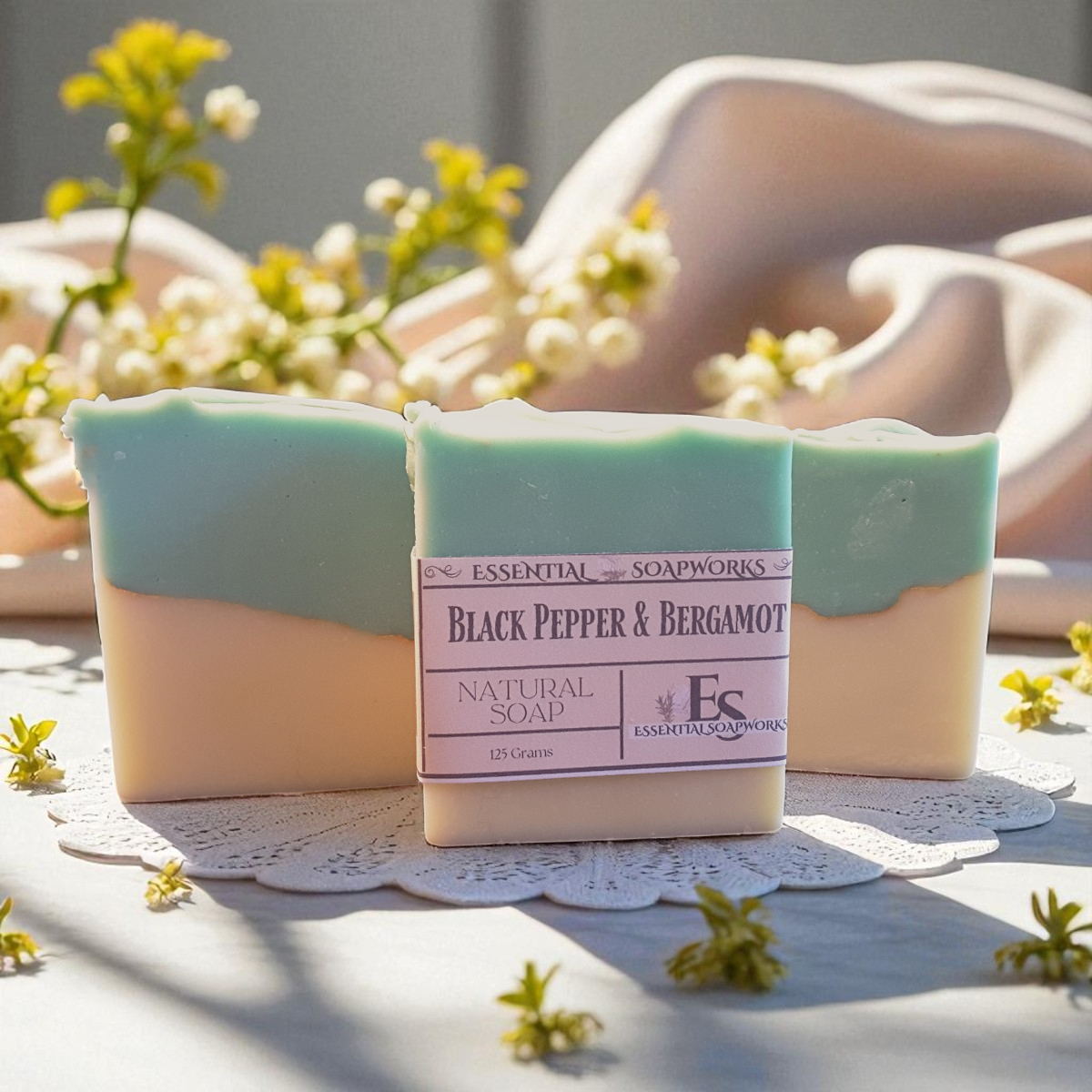 Two bars of Black Pepper & Bergamot soap on a white doily with flowers in the background.