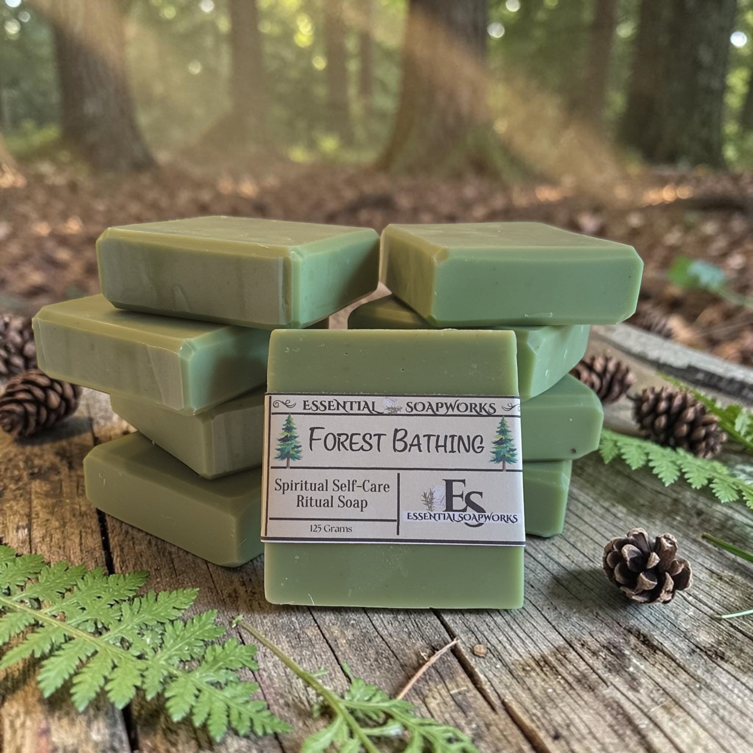 Five bars of sage green ritual soap stacked on a rustic wooden surface outdoors, with pinecones and green leaves in the foreground and a blurred forest background. The front bar has a white label reading 'ESSENTIN SOAPWORKS Forest Bathing Spiritual Self-Care Ritual Soap'.