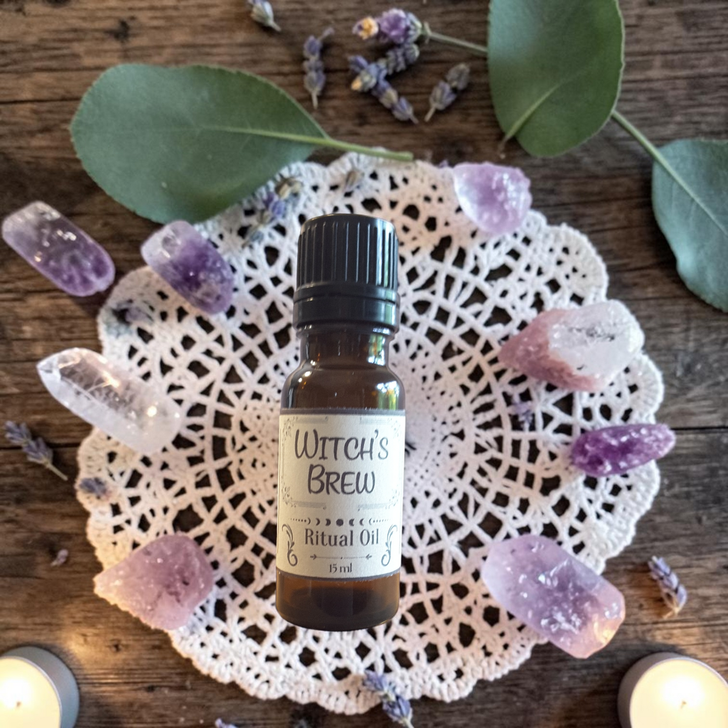 A small amber glass bottle labeled 'Witch's Brew Ritual Oil' sits centered on a white lace doily, surrounded by purple crystals, dried lavender, green leaves, and candles on a rustic wooden surface.
