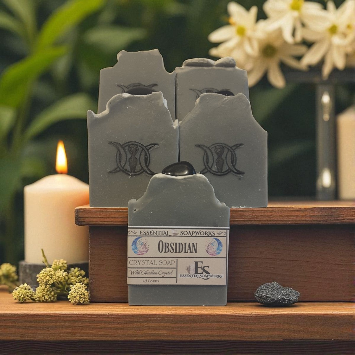 Obsidian crystal soap with a triple goddess symbol on a wooden shelf with a candle and flowers in the background