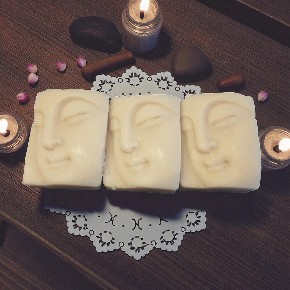 Three soap bars with buddha facial designs on a wooden surface with candles and decorative elements.