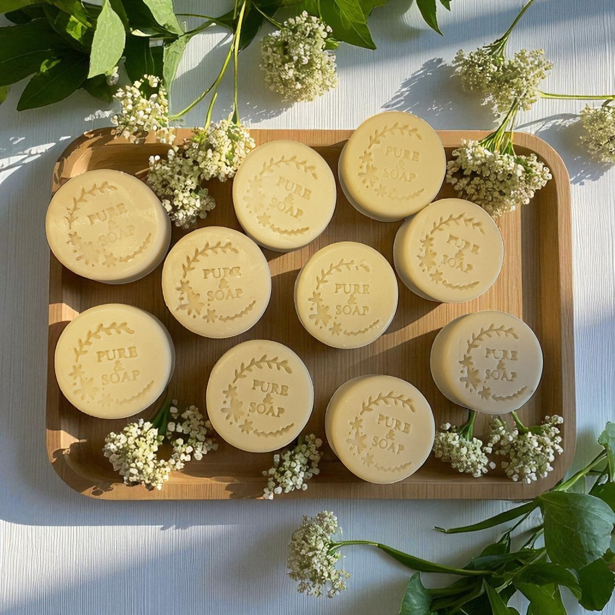 Wooden tray with soap bars engraved with 'PURE SOAP' surrounded by white flowers on a light background
