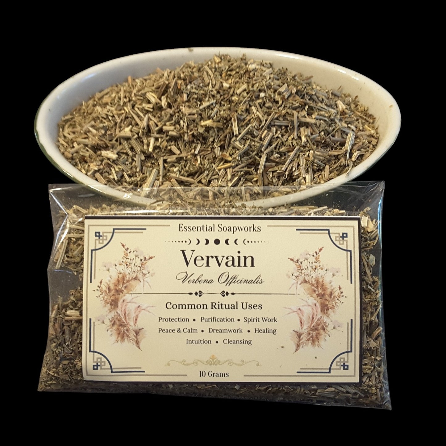 A clear plastic bag of Vervain Herb for witchcraft with a cream label, shown in front of a white bowl filled with dried vervain herb pieces on a black background.