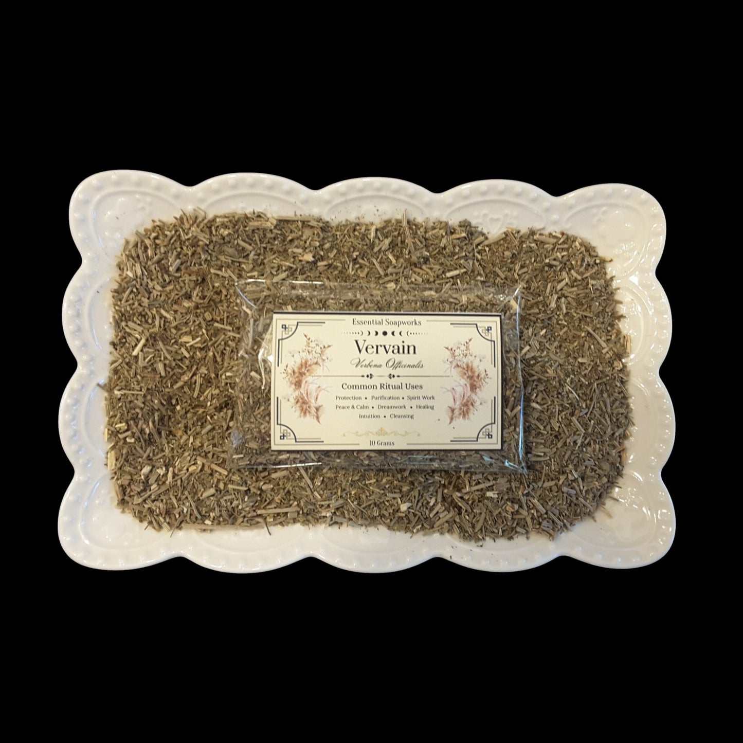 A square white ceramic dish with a scalloped edge holds loose dried vervain for witchcraft surrounding a clear plastic bag with a label that reads 'Vervain'. The product is photographed from above against a solid black background.