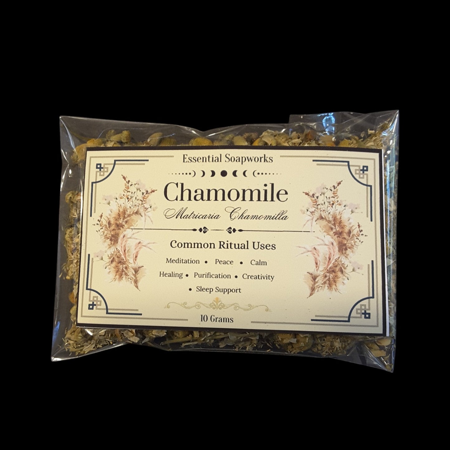 A 10-gram clear plastic bag of chamomile flowers by Essential Soapworks, featuring a cream-colored label with botanical illustrations and text, set against a solid black background.