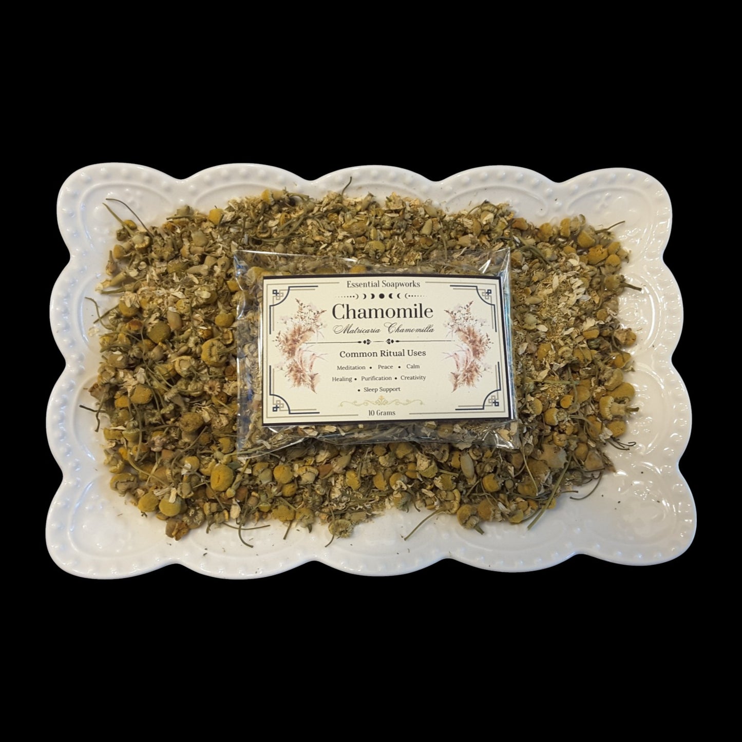 A clear plastic packet of chamomile flowers sits centered on a white scalloped ceramic plate filled with dried chamomile flowers, photographed against a solid black background. The packet has a decorative white label reading 'Chamomile' and 'Common Ritual Uses.