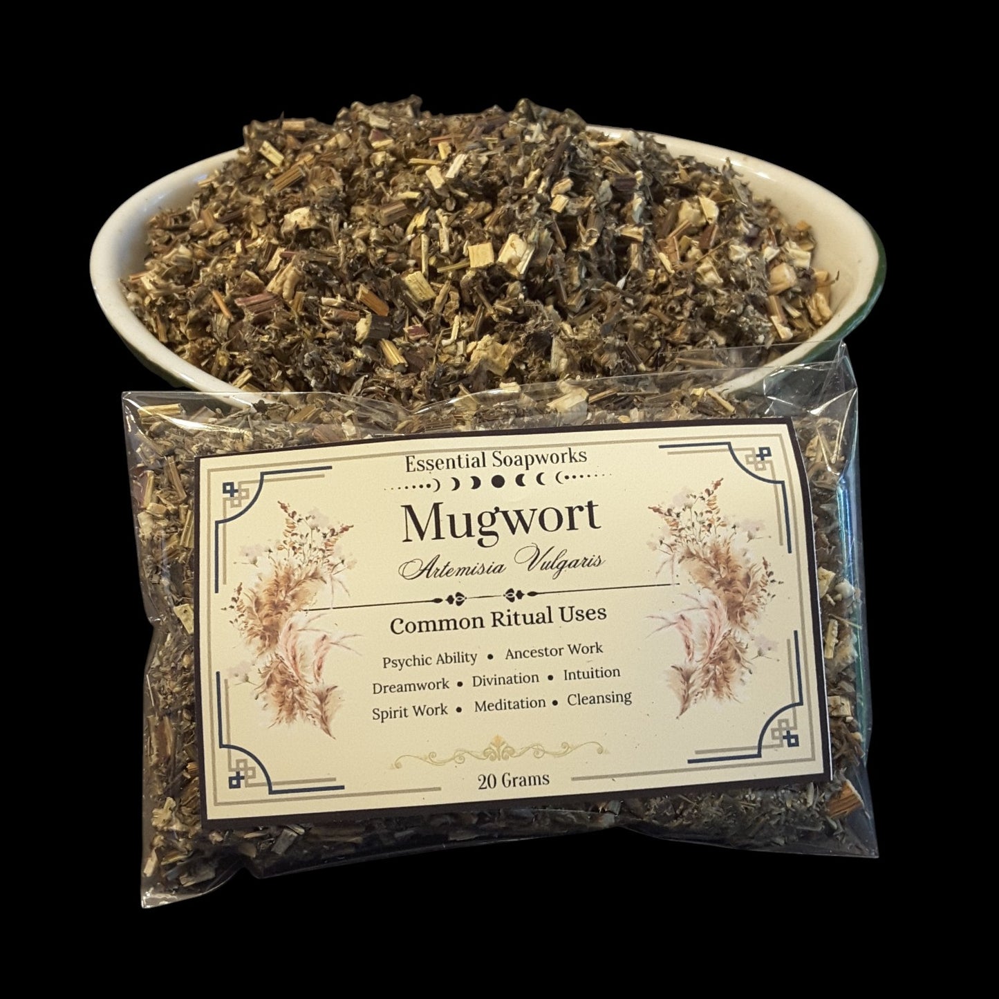 A clear plastic bag of mugwort herb with a cream label reading 'Mugwort Alchemica Vulgaris' and 'Essential Soapworks,' placed in front of a white bowl filled with loose dried mugwort on a black background.