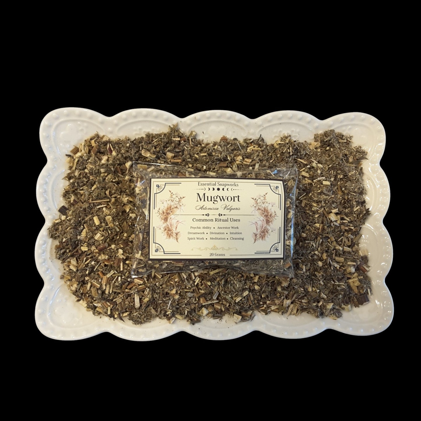 A square white ceramic plate with a scalloped edge holds a clear plastic bag of loose mugwort herb. The herb is a dry, textured blend of green and brown herb pieces, and a cream-colored label with black text and botanical illustrations is centered on the package.