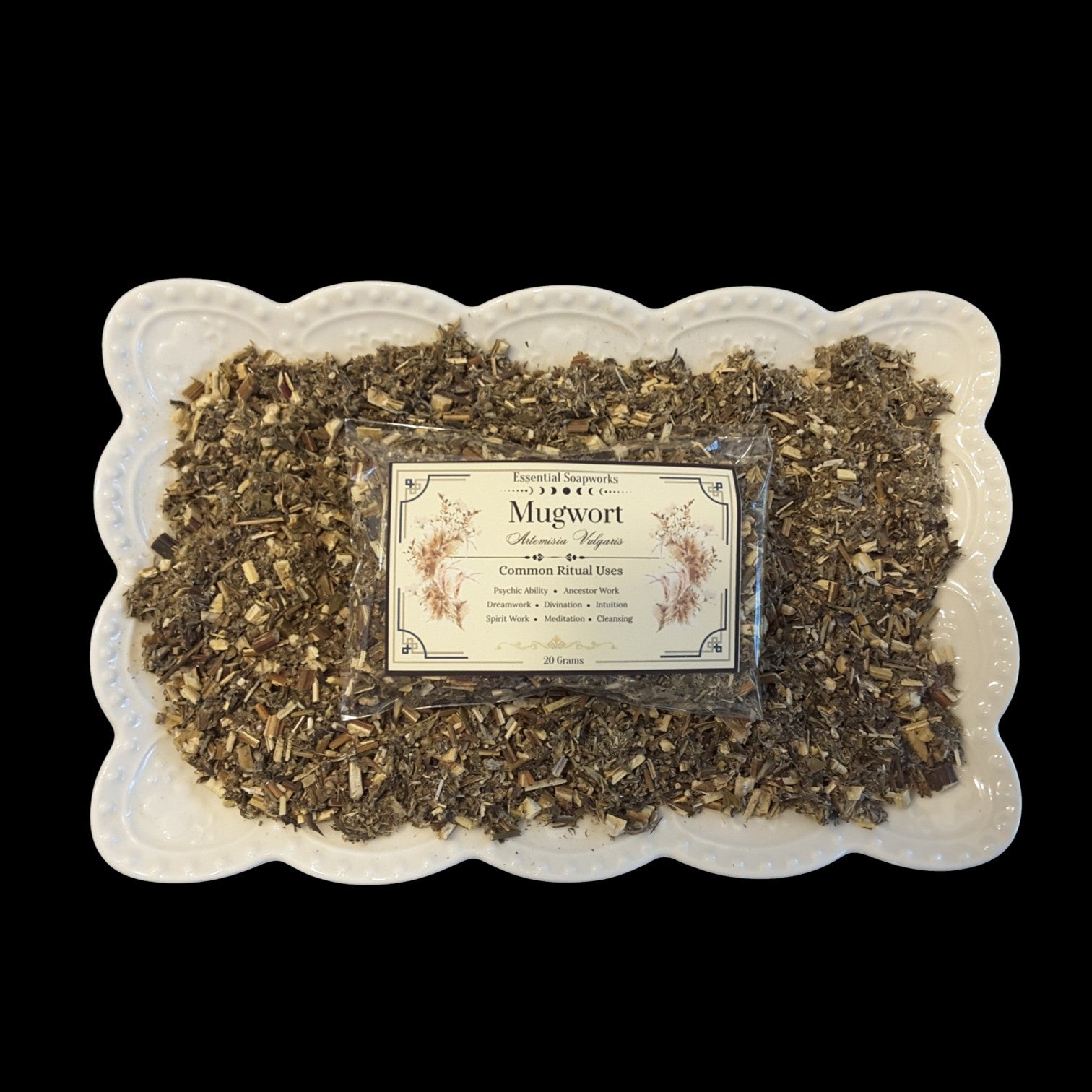 A square white ceramic plate with a scalloped edge holds a clear plastic bag of loose mugwort herb. The herb is a dry, textured blend of green and brown herb pieces, and a cream-colored label with black text and botanical illustrations is centered on the package.