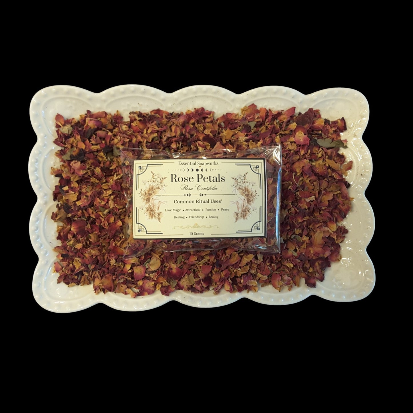 A square white scalloped dish filled with dried rose petals around a small clear plastic package labeled 'Rose Petals' shown on a black background.