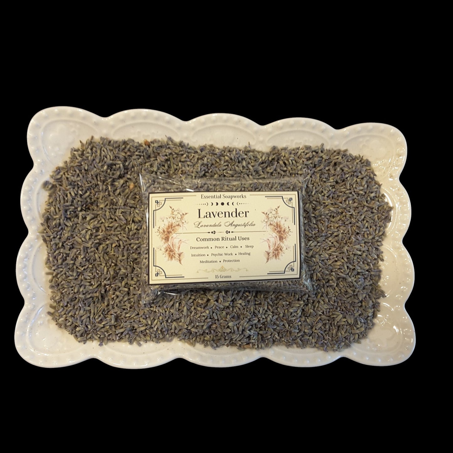 A packaged bundle of lavender buds arranged on a white scalloped ceramic plate against a solid black background. The center of the bundle contains a clear plastic bag with a cream-colored label that reads 'Lavender'