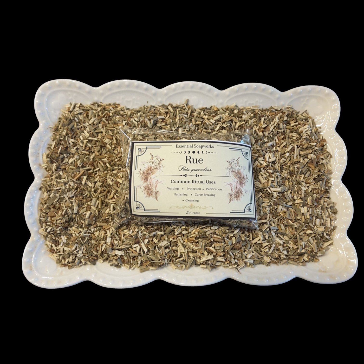 A square white ceramic dish with a scalloped edge holds loose dried rue with a clear plastic bag and printed label centered on top. The label reads 'Rue'.