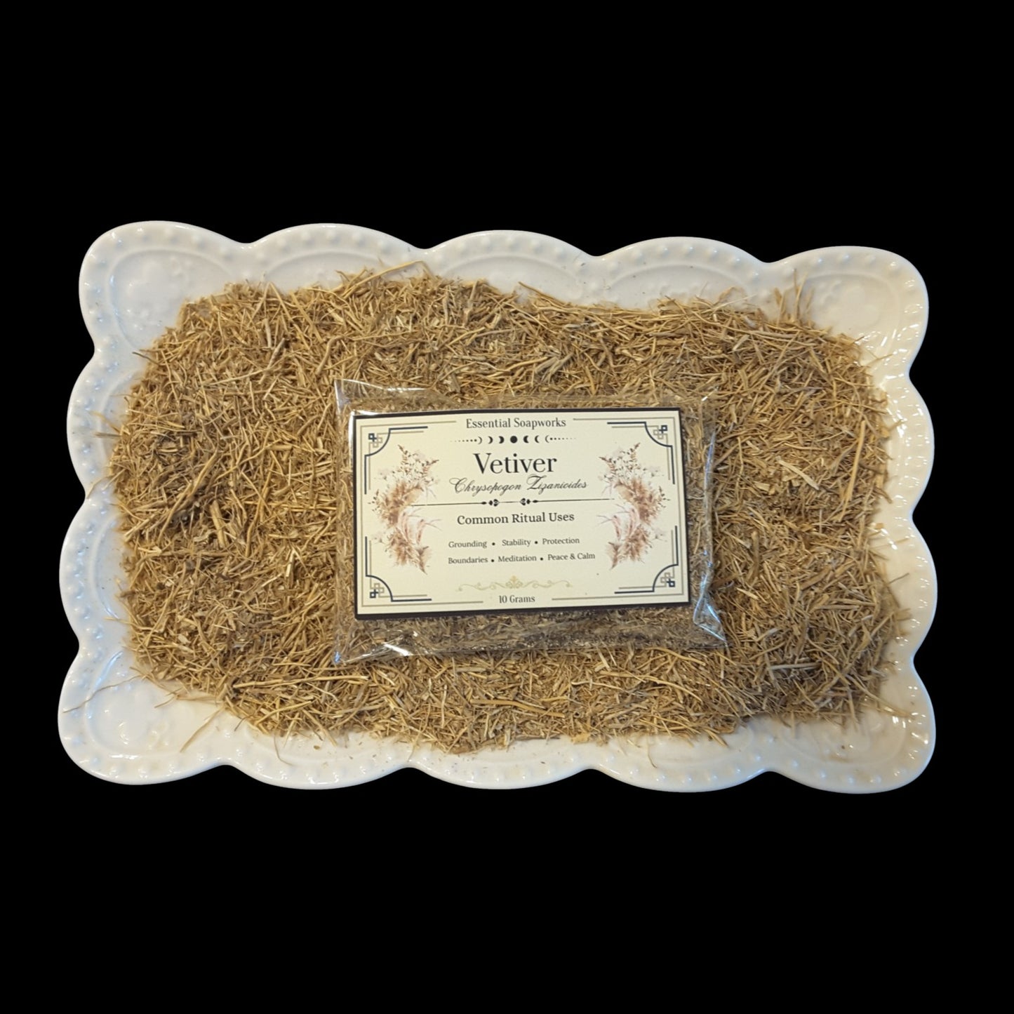 A clear plastic bag of Vetiver root wrapped in a clear bag with a decorative cream label, placed on top of loose golden-brown vetiver roots inside a white scalloped ceramic dish, against a solid black background.