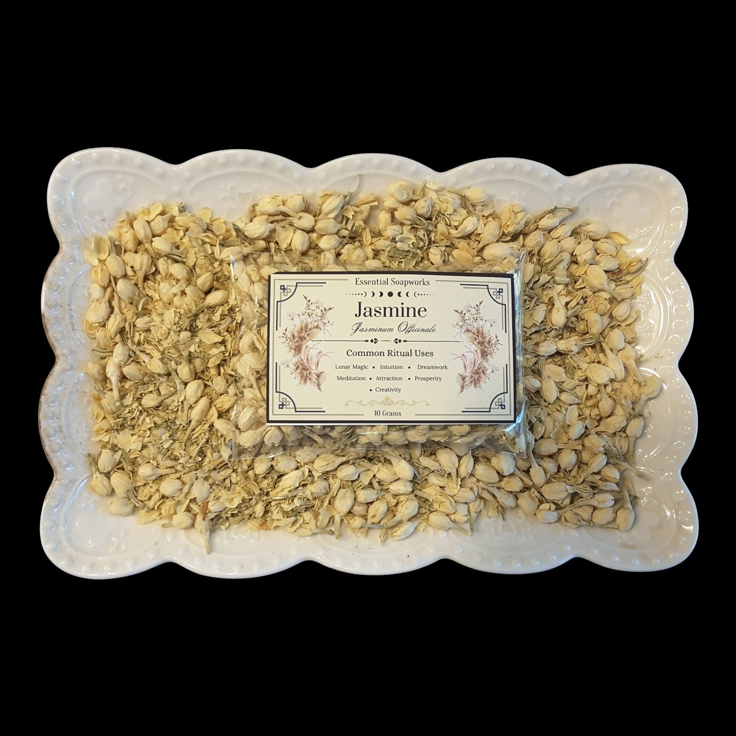 A white scalloped ceramic dish filled with dried jasmine flowers and a central product label reading 'Jasmine' and 'Essential Oils,' photographed against a solid black background.