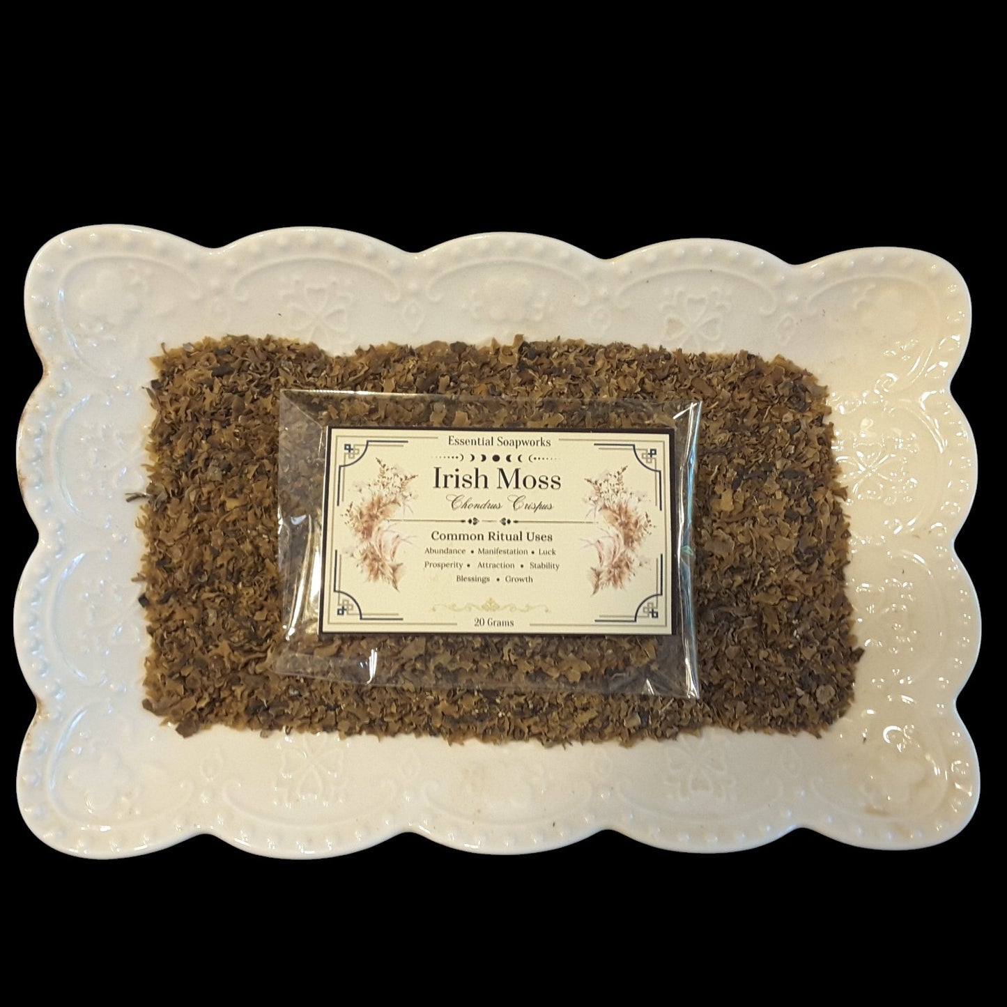 A package of Irish Moss botanicals placed on top of loose dried Irish Moss inside a clear plastic bag, set on a white scalloped ceramic platter against a solid black background.