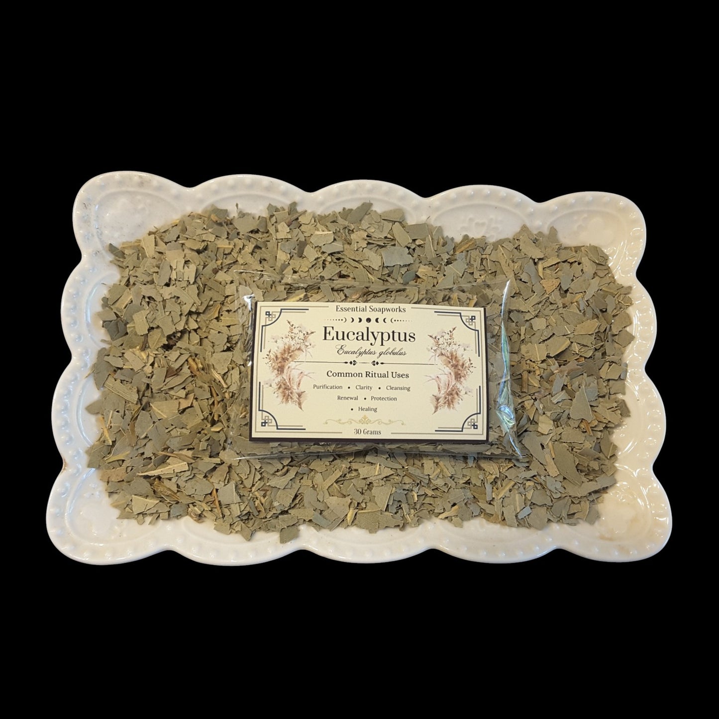 A package of Eucalyptus sits in the center of a white scalloped ceramic dish filled with loose dried eucalyptus leaves, set against a solid black background.