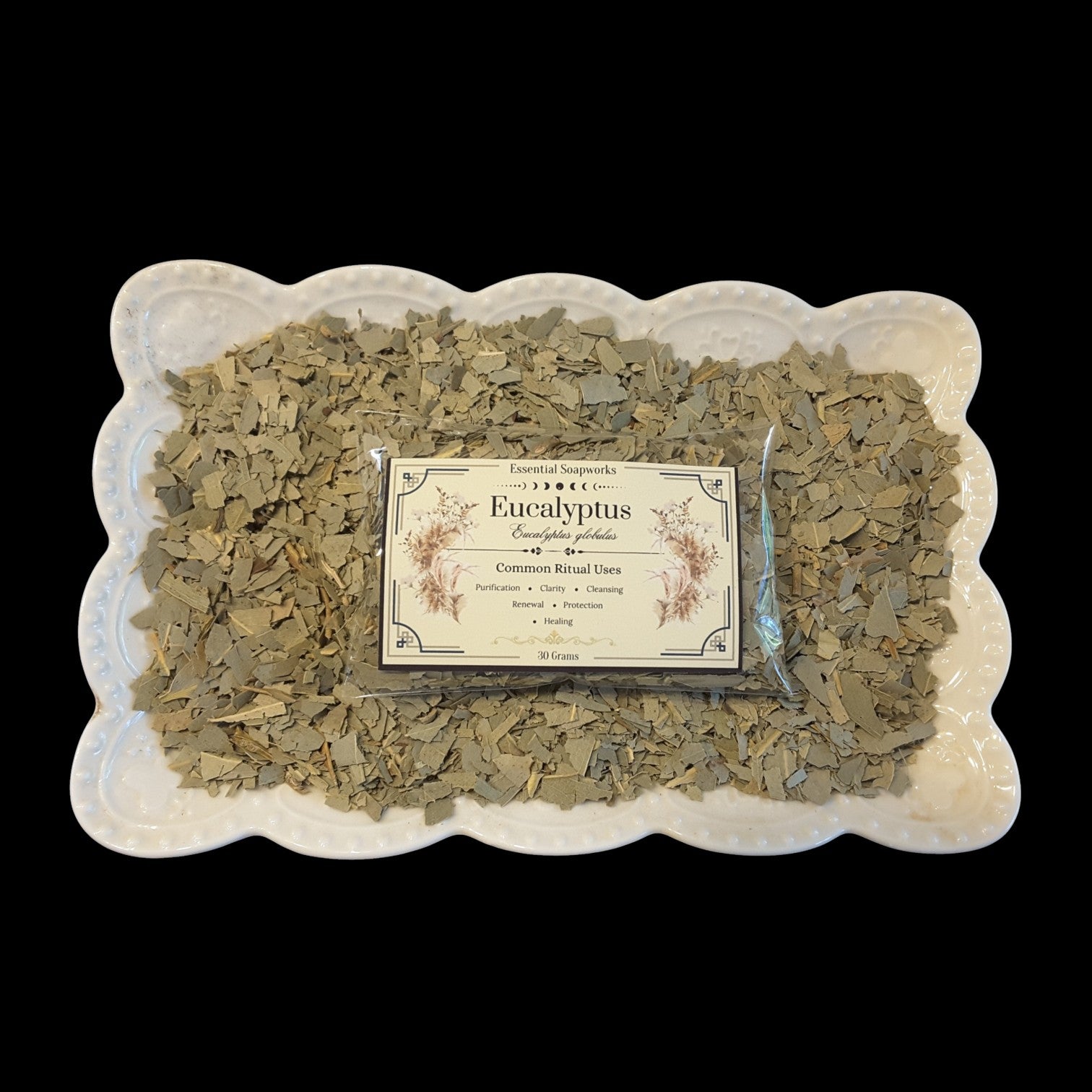 A package of Eucalyptus sits in the center of a white scalloped ceramic dish filled with loose dried eucalyptus leaves, set against a solid black background.