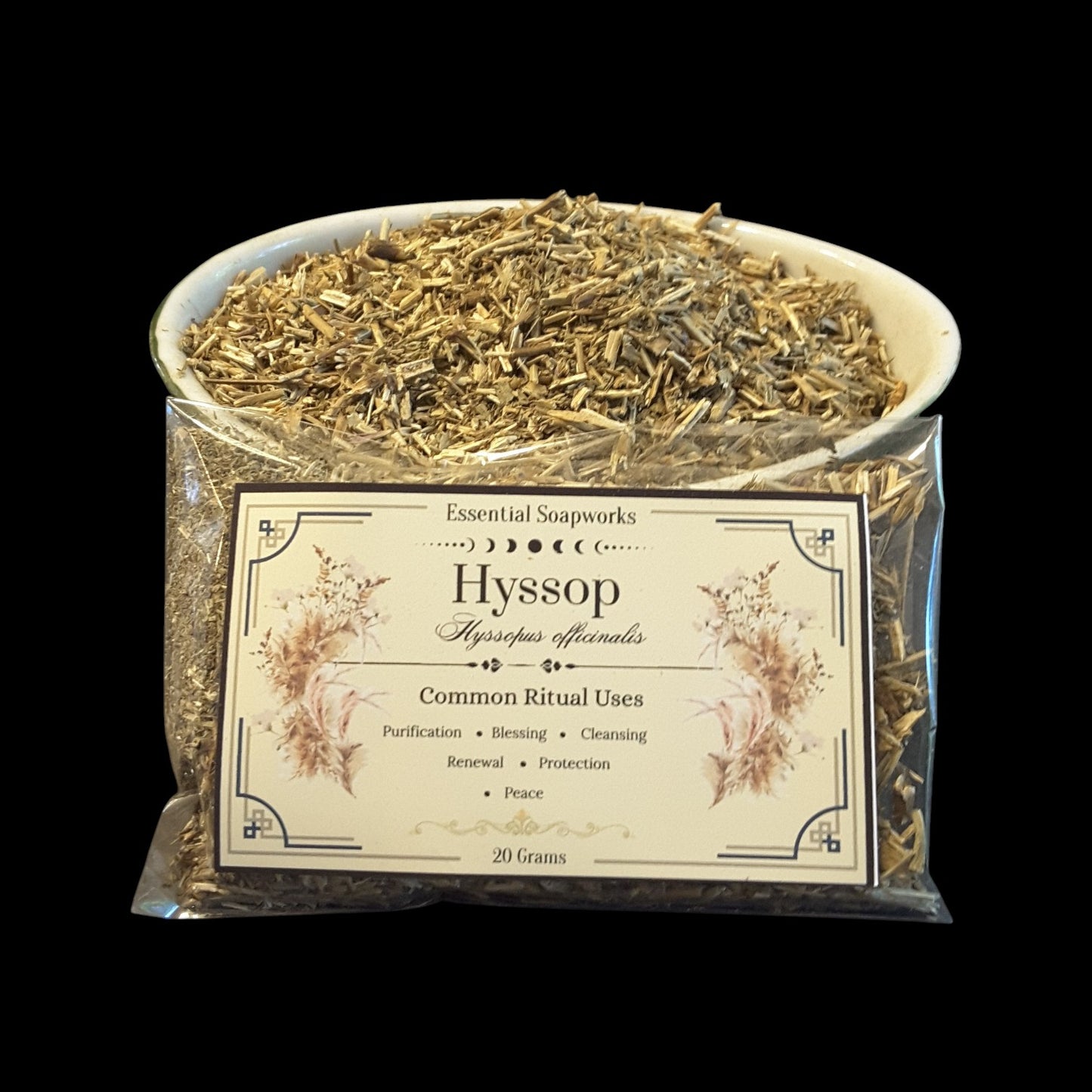 A clear plastic bag filled with dried hyssop herb, with a white bowl filled with the same dried herb on top, and a cream-colored label on the front. The label reads 'Essential Soapworks Hyssop' and 'Common Ritual Uses'.
