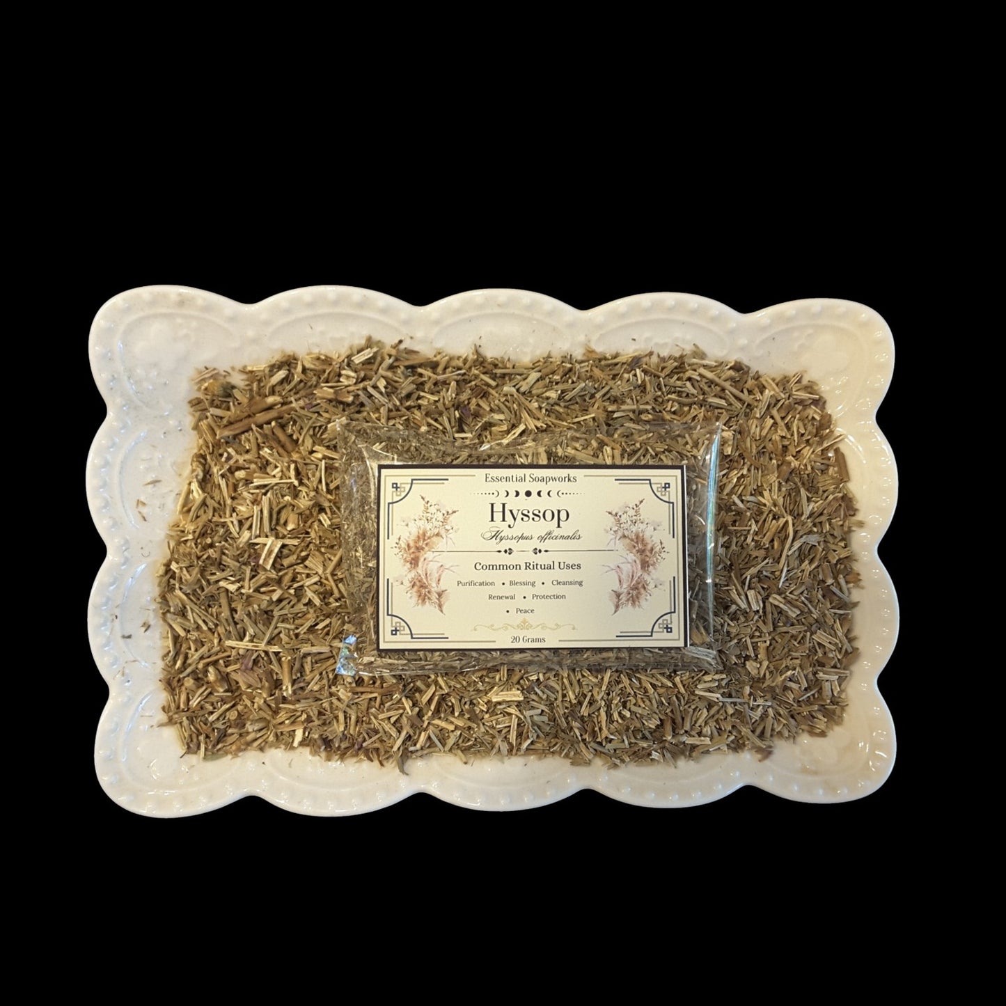 A top-down view of dried hyssop botanicals in a clear plastic bag with a vintage-style cream label, presented on a white scalloped ceramic dish against a solid black background.
