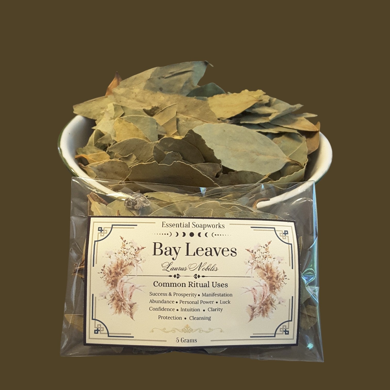 A clear plastic bag containing dried bay leaves, with a white bowl filled with more dried leaves behind it. A label on the front reads 'Essential Soapworks Bay Leaves' and lists common ritual uses.