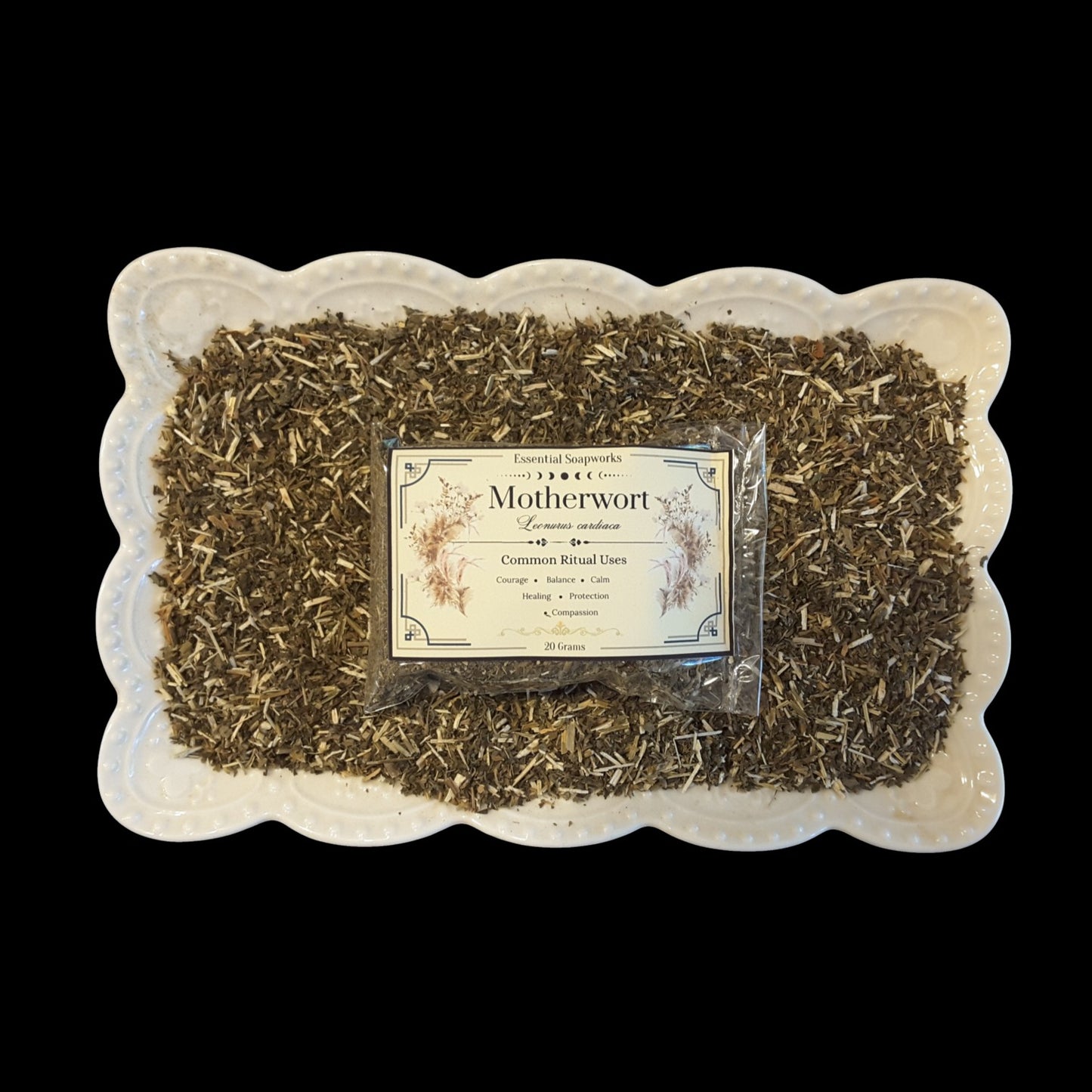 A top-down view of a white scalloped ceramic dish filled with dried motherwort, with a clear plastic bag of loose-leaf motherwort centered on top and a vintage-style label reading 'Motherwort' in the middle, set against a solid black background.