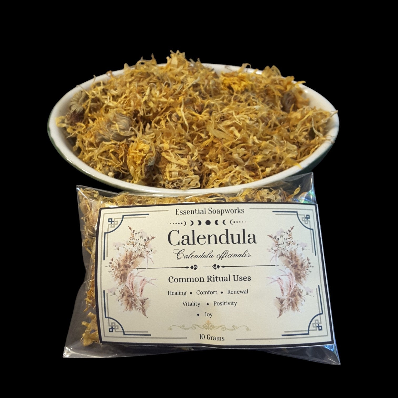 A clear plastic bag of Calendula flowers with a cream label reading 'Essential Soapworks Calendula' sits in front of a white bowl filled with dried golden-yellow calendula petals, all photographed against a solid black background.