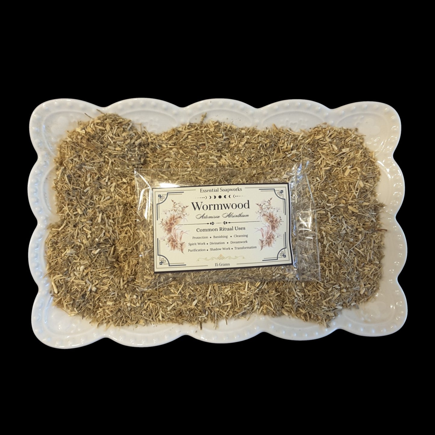 A clear plastic bag of Wormwood for witchcraft, placed on top of dried wormwood leaves inside a white scalloped dish, against a solid black background.