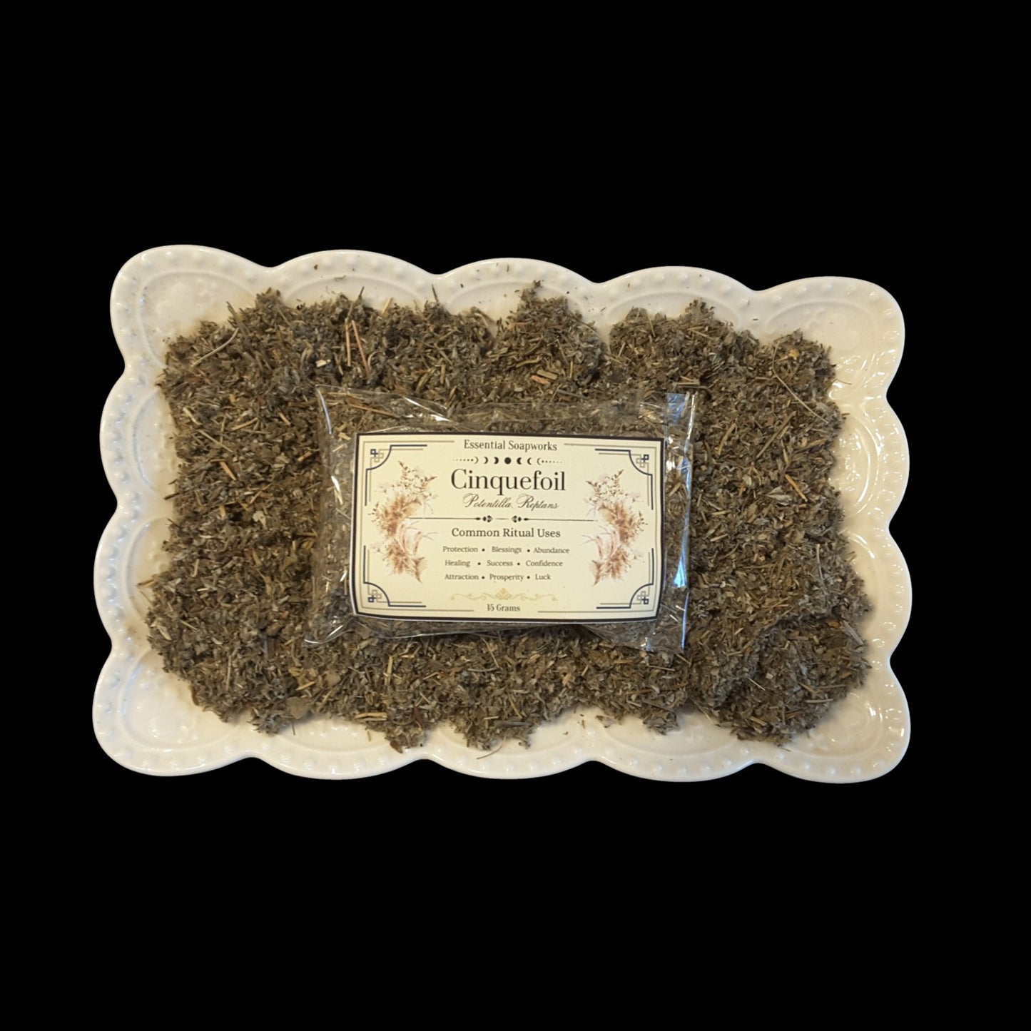 A package labeled 'Cinquefoil' sits on top of loose dried herbs arranged in a white scalloped dish against a solid black background.