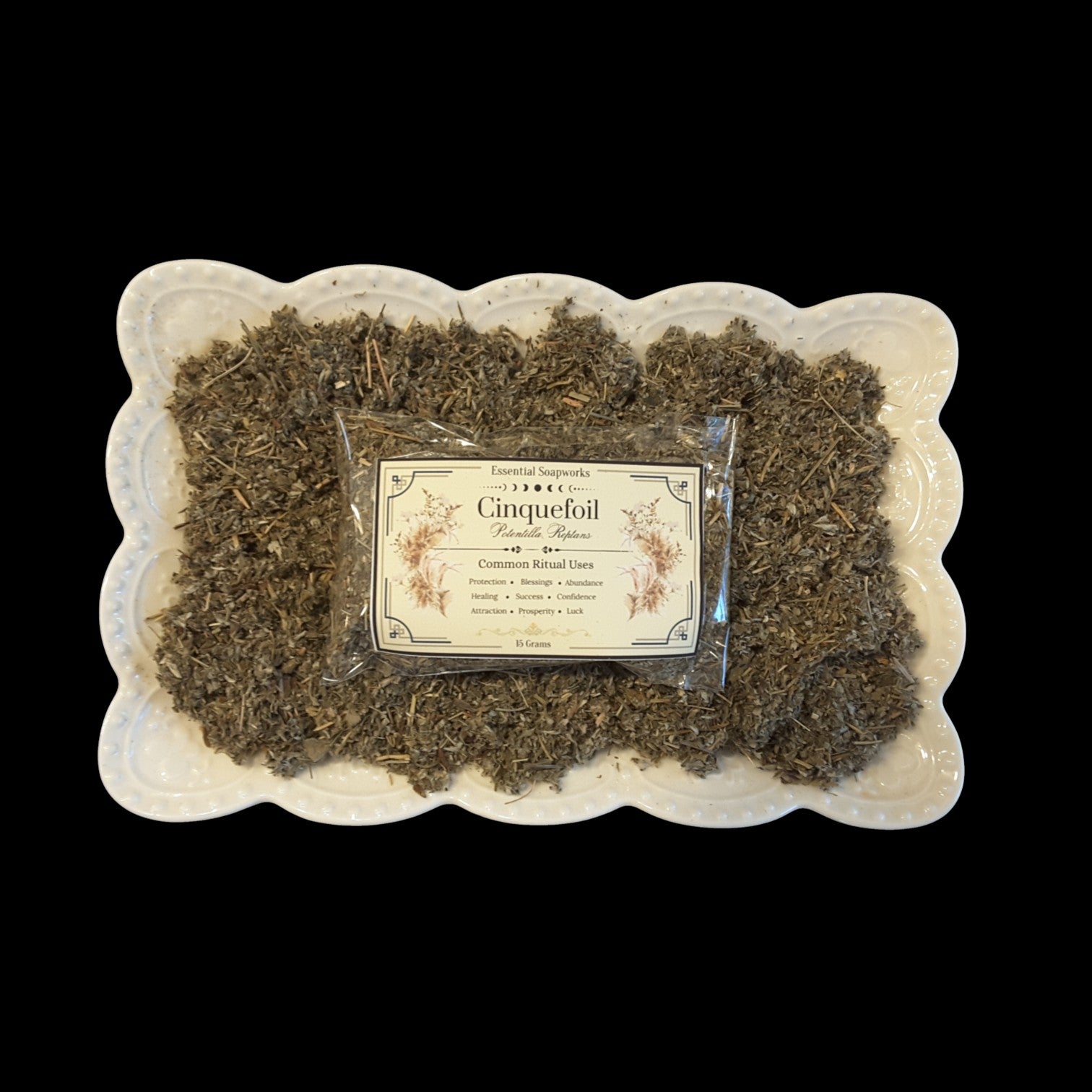 A package labeled 'Cinquefoil' sits on top of loose dried herbs arranged in a white scalloped dish against a solid black background.