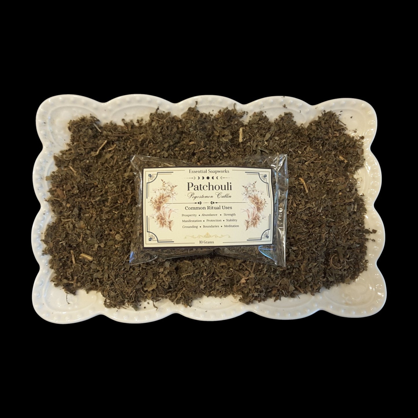 A small clear packet labeled 'Patchouli' with a cream label sits centered on a bed of dried brown patchouli leaves in a white scalloped dish, photographed against a black background.