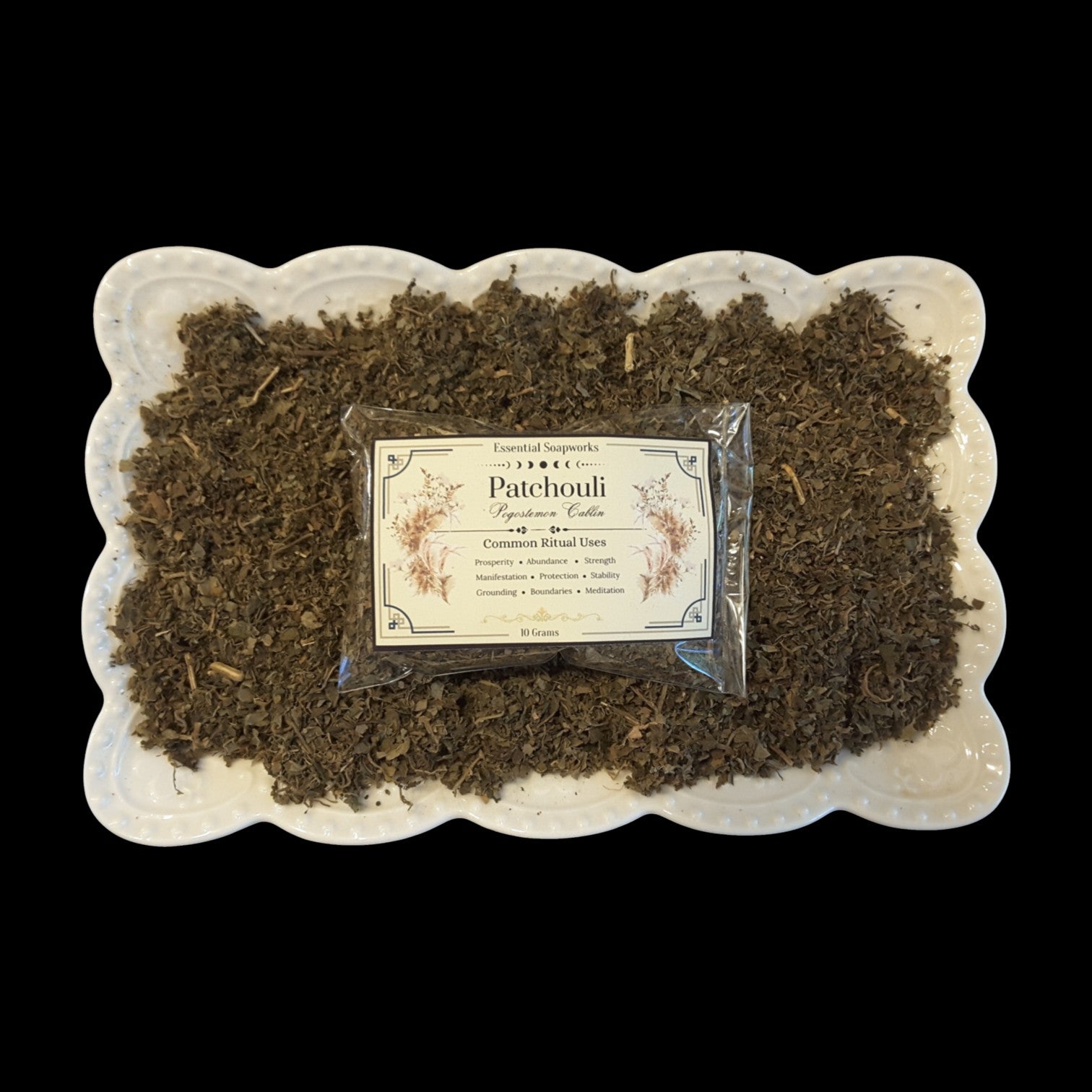 A small clear packet labeled 'Patchouli' with a cream label sits centered on a bed of dried brown patchouli leaves in a white scalloped dish, photographed against a black background.