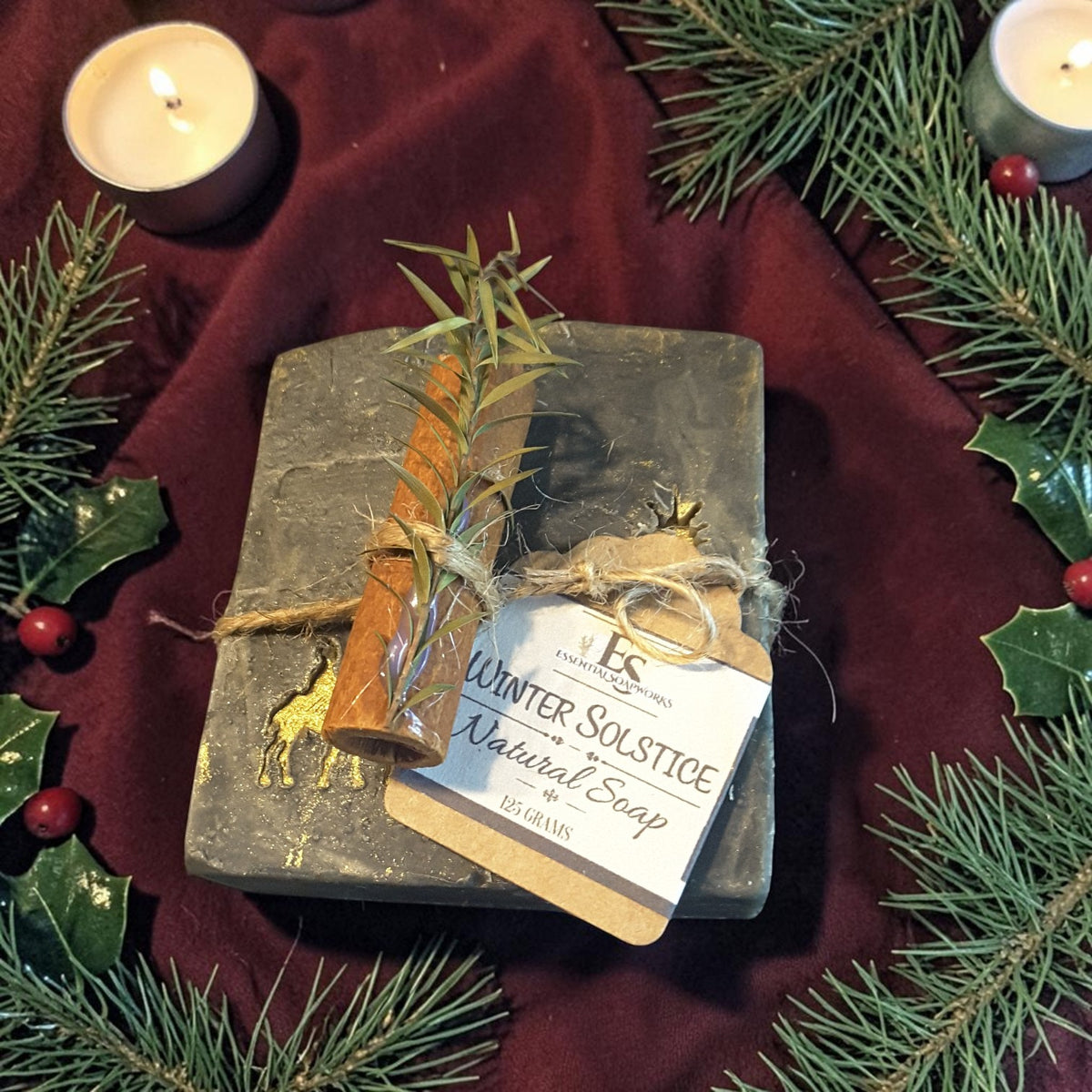 winter solstice soap tied with twine and a cinnamon stick along with greenery on a red fabric surface with pine needles and candles