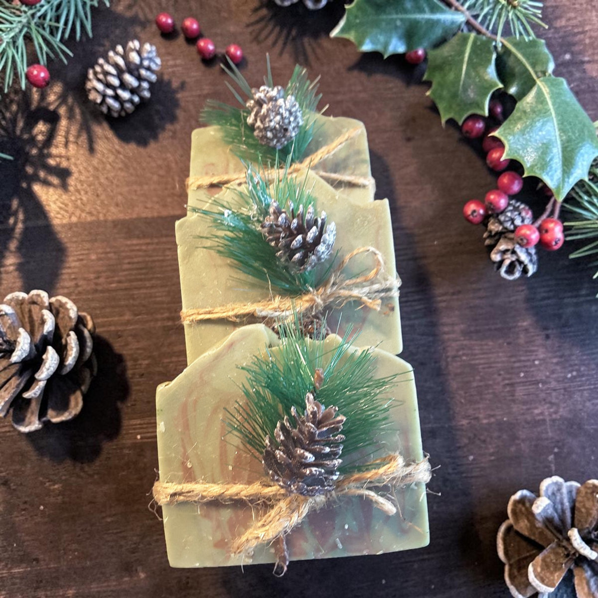 three bars of alpine frost christmas soap on a wooden surface with pine cones and holly