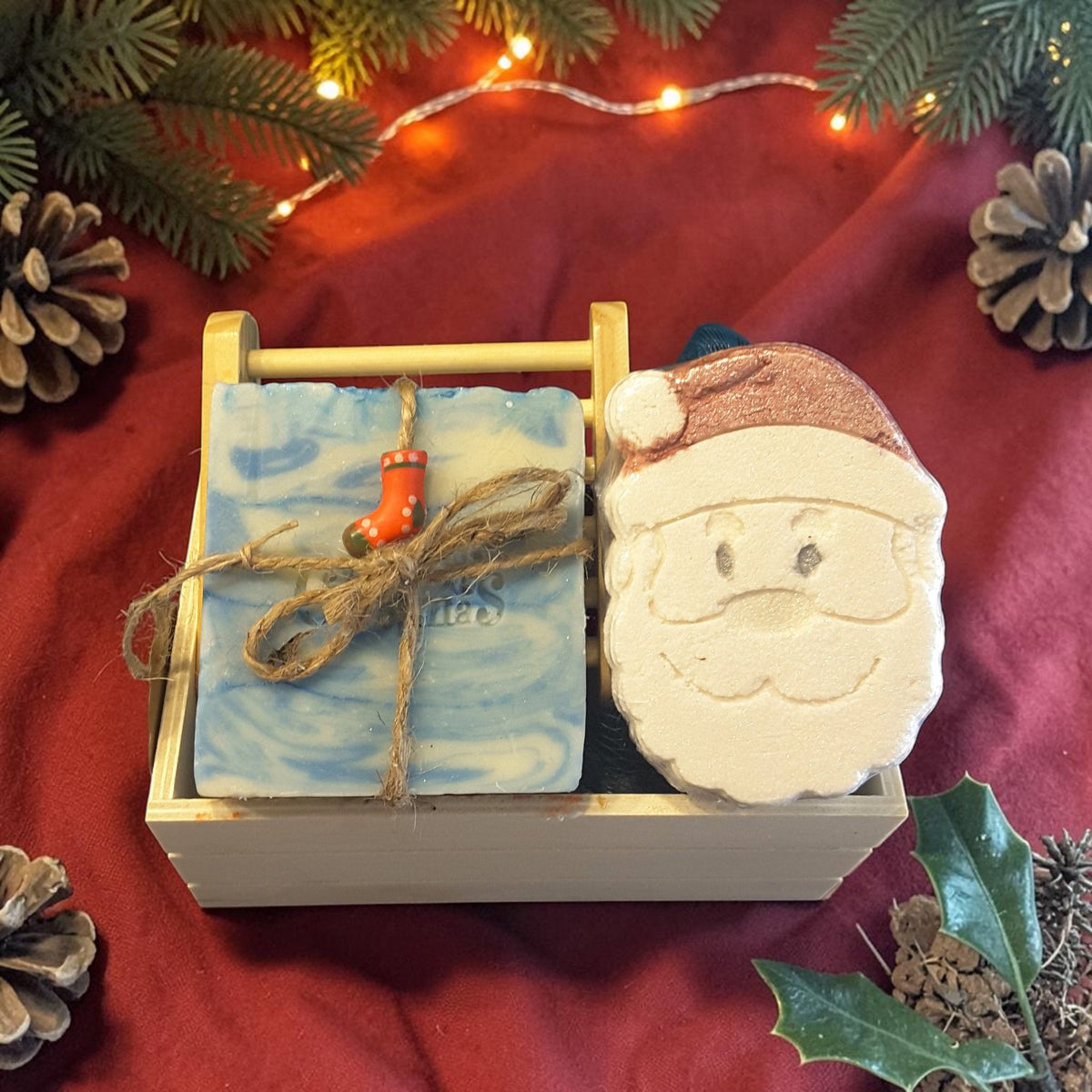 snowdrift christmas gift crate spa set featuring snowdrift soap, santa shaped bath bomb, bamboo soap ladder and loofah on a red fabric surface with pine cones and lights 