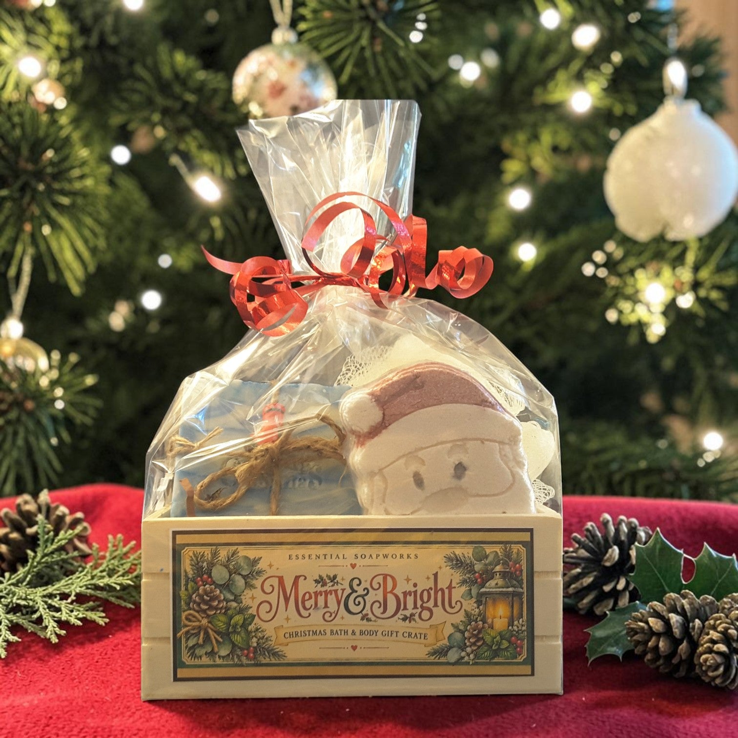 snowdrift christmas gift crate spa set featuring snowdrift soap, santa shaped bath bomb, bamboo soap ladder and loofah gift wrapped in a wooden crate on a red fabric surface with a christmas tree in the background