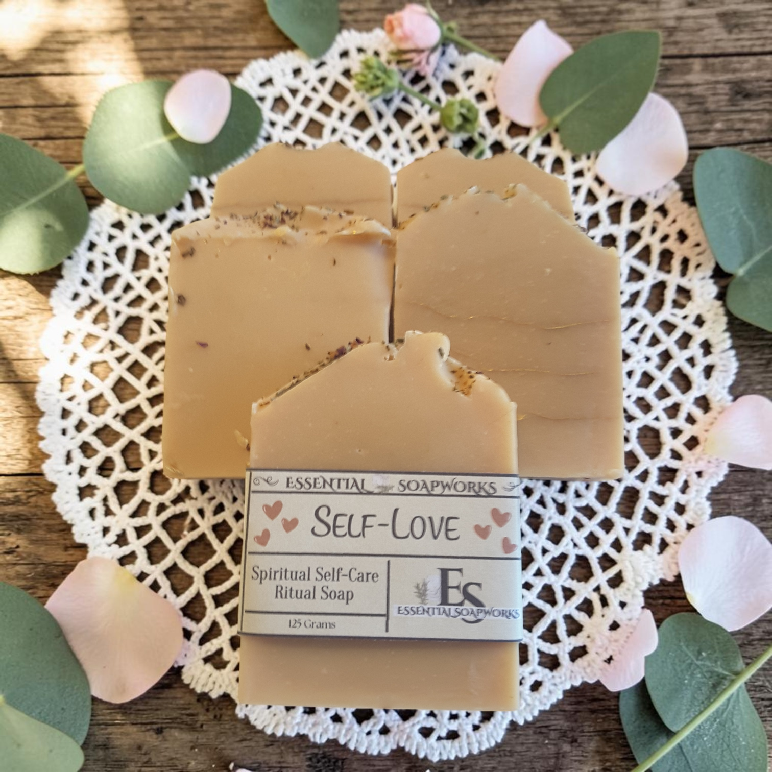 Three rectangular bars of tan ritual soap with visible botanical inclusions, arranged on a white lace doily over a rustic wooden surface with green eucalyptus leaves and pale pink petals. The front bar has a paper label reading 'ESSENTIAL SOAPWORKS SELF-LOVE Spiritual Self-Care Ritual Soap'.