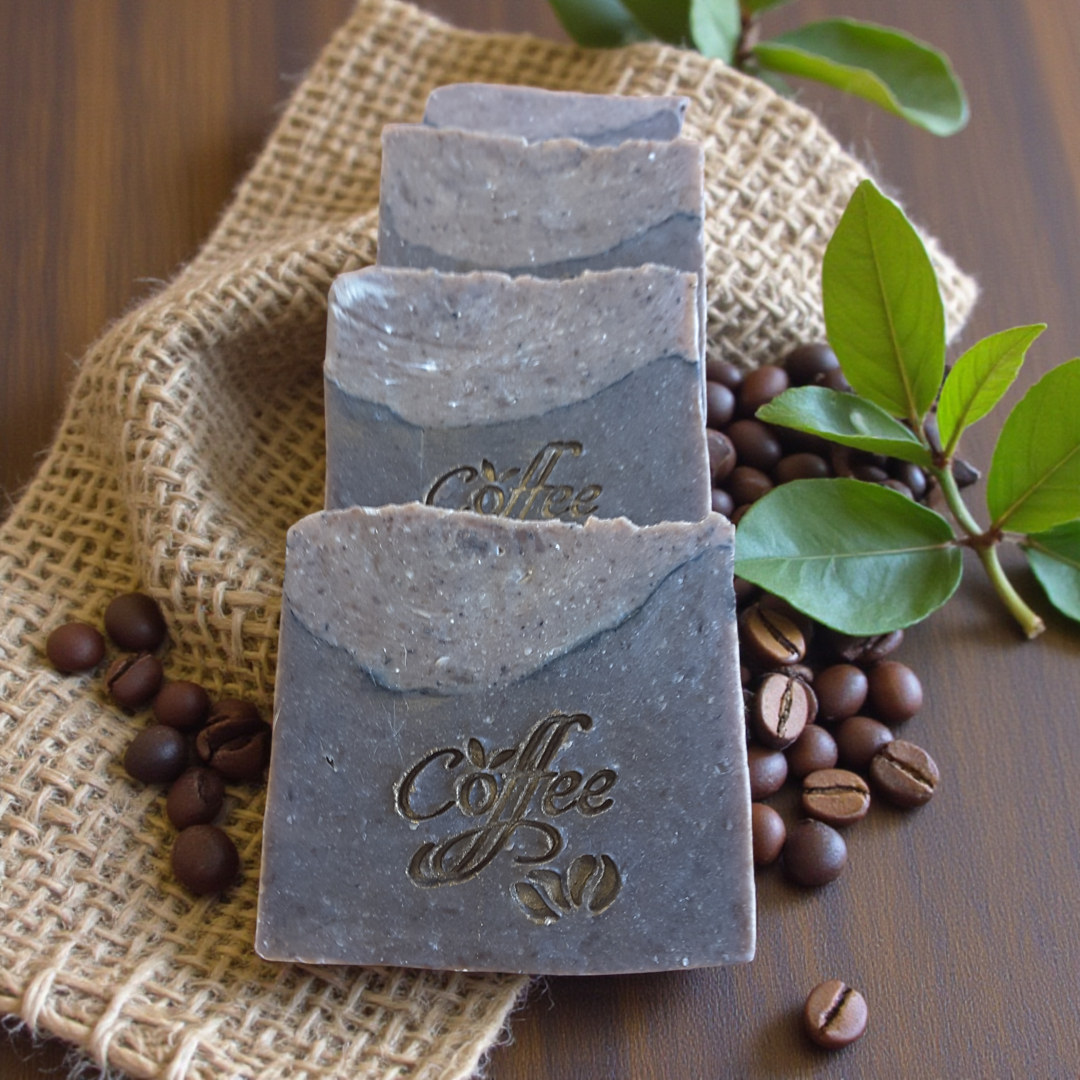 Three bars of coffee-scented soap on a rustic surface with coffee beans and leaves.
