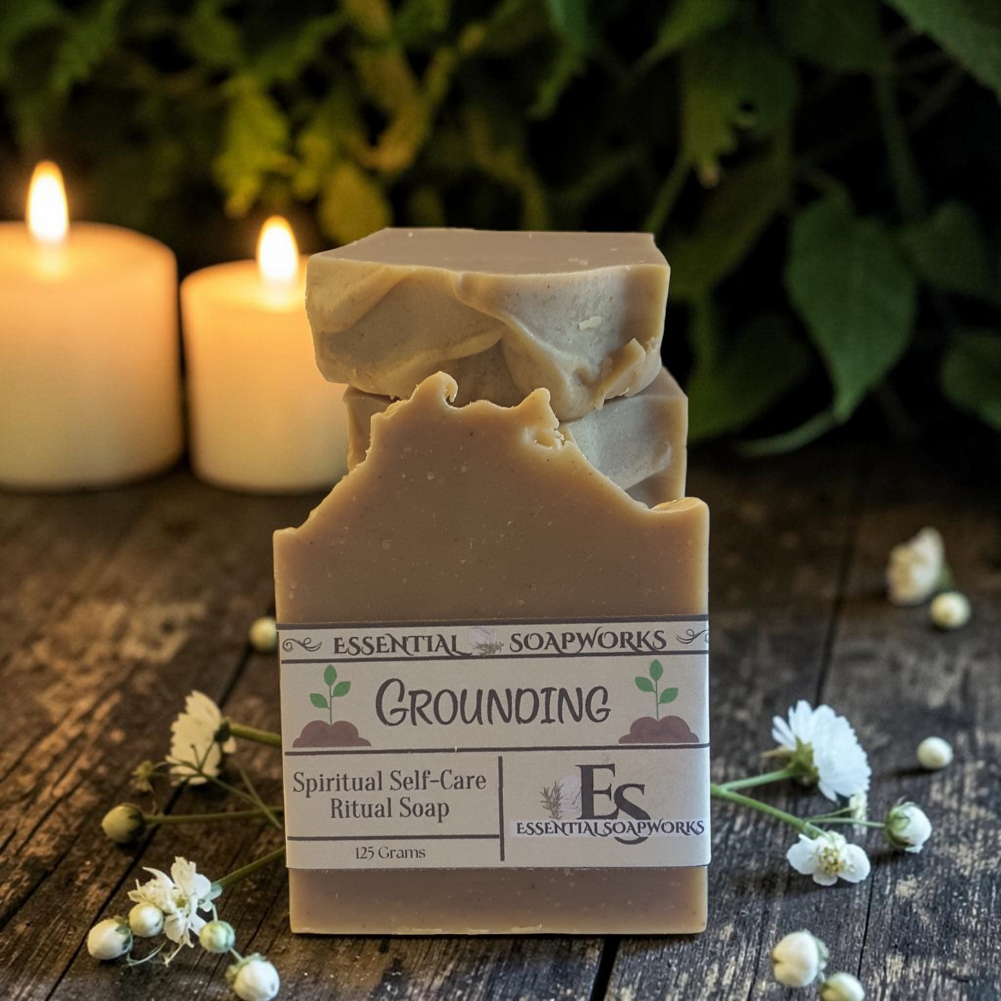 A handmade bar of soap labeled 'Grounding' by Essential Soapworks, styled on a rustic wooden surface with white flowers and glowing candles in the background.