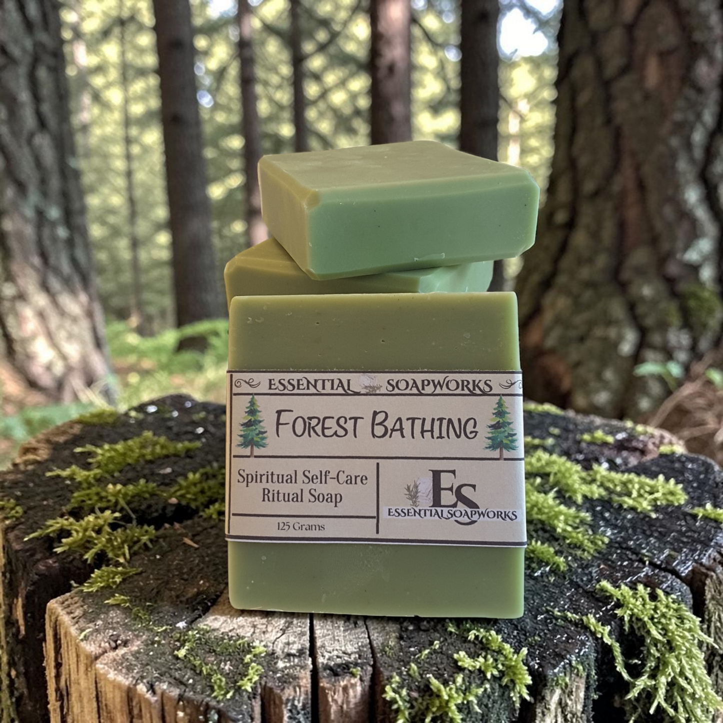 Three bars of green ritual soap stacked on a mossy tree stump in a forest setting. The front bar has a white paper label that reads 'ESSENTIAL SOAPWORKS Forest Bathing Spiritual Self-Care Ritual Soap'.