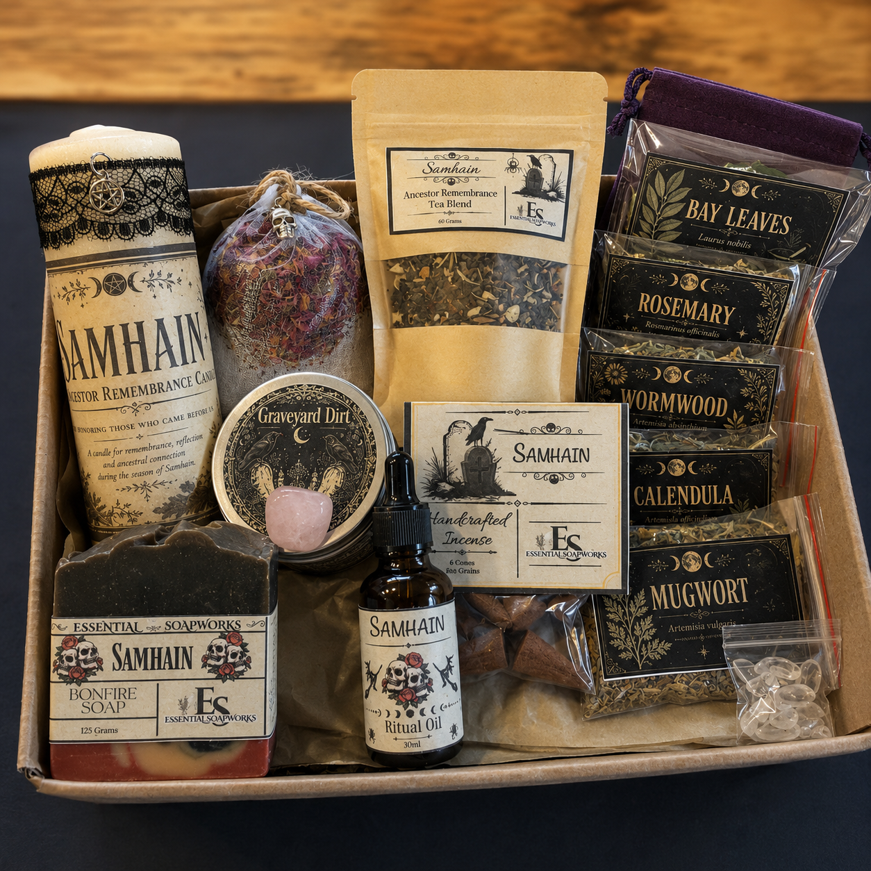 A Samhain ritual kit filled with various natural products including soap, ritual oil, tea, dried flowers, and herbal bundles, arranged in a cardboard tray on a dark surface.
