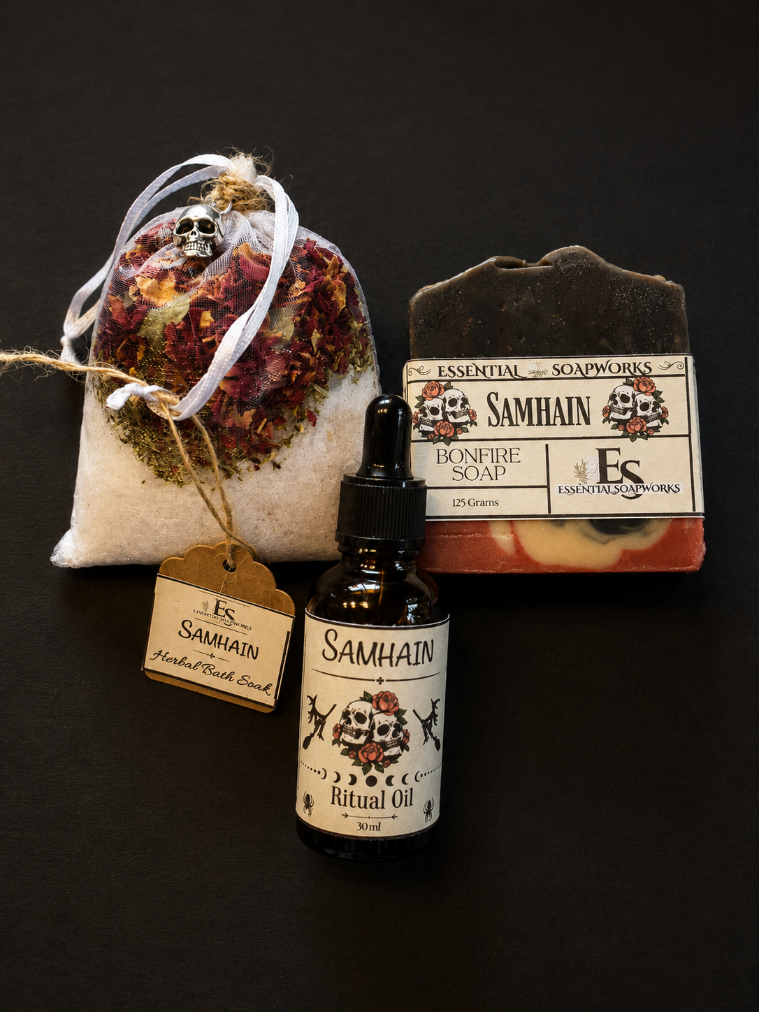 A Samhain ritual kit featuring a bottle of Samhain Ritual Oil, a bar of Bonfire Soap, a dried flower sachet with a skull ornament, and a tag, all arranged on a dark background.