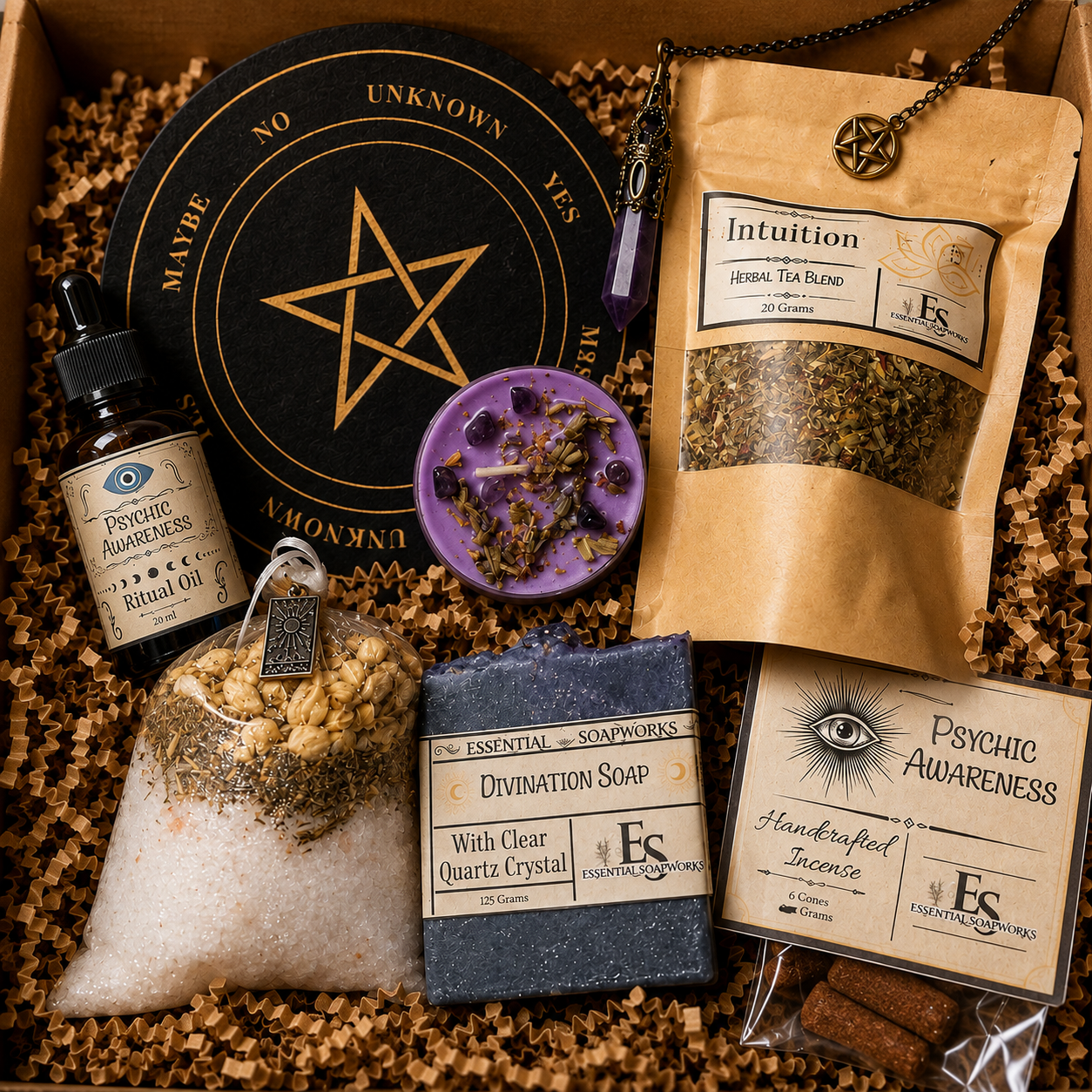 A curated spiritual gift box or product assortment arranged in crinkle paper inside a box. Visible items include a round black tarot card with a gold pentagram, a purple wax candle with dried flowers, a pouch labeled 'Intuition Herbal Tea Blend,' a small bottle labeled 'Psychic Awareness Ritual Oil,' a bag labeled 'Divination Soap,' a bar of soap labeled 'Essential Soaps Divination Soap,' a small card labeled 'Psychic Awareness,' and a necklace with a pentagram pendant.