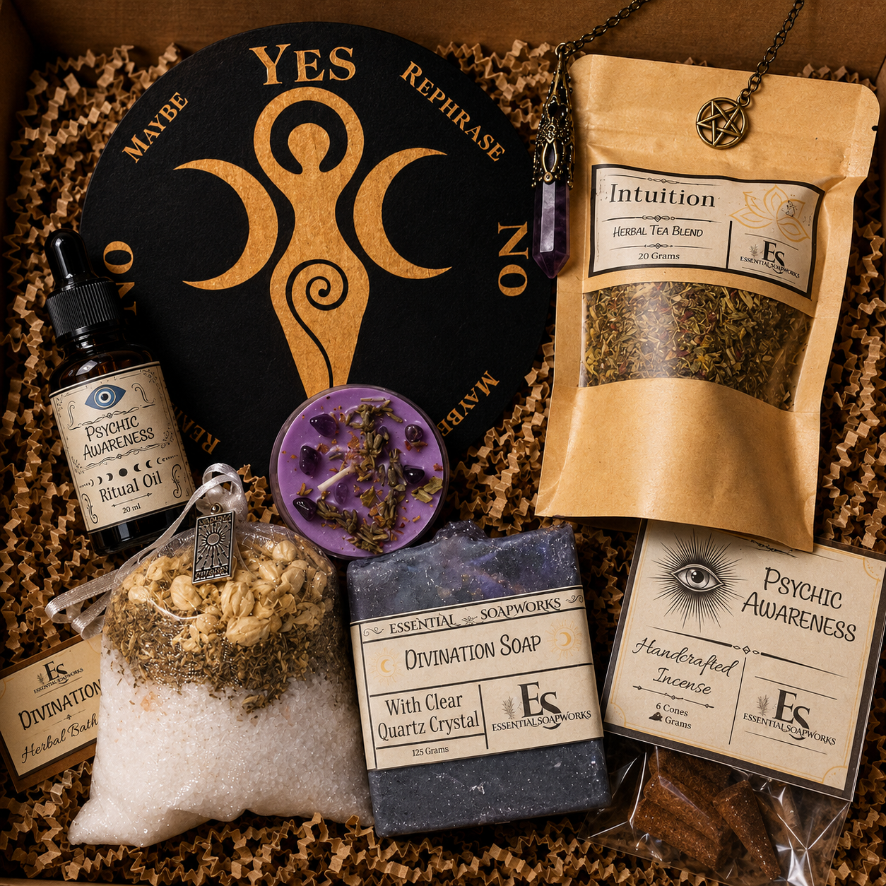 A curated spiritual gift box or product bundle arranged in a box with brown crinkle paper filler. Visible items include a black circular sign with gold moon phases and the words YES/NO/MAYBE, a kraft pouch labeled Intuition, a small bottle labeled Sacred Tea Blend, a round purple soap bar, a bag labeled Divination Salt, a dark gray soap bar labeled Divination Soap, a card labeled Psychic Awareness Handcrafted Incense, and a necklace with a pentagram pendant.