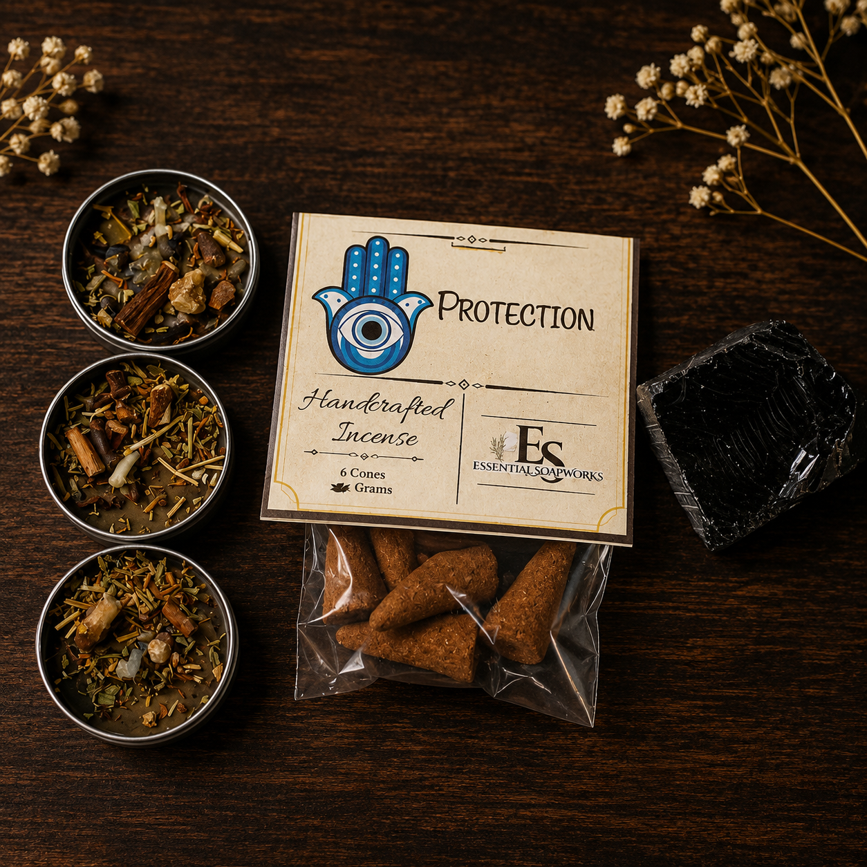 A styled flat-lay of a handcrafted incense set on a dark wood background. The set includes a kraft paper box labeled 'PROTECTION' with a blue Hamsa hand logo, a clear bag of three brown incense cones, three small metal tins filled with dried botanical incense blends, and a black rectangular box.