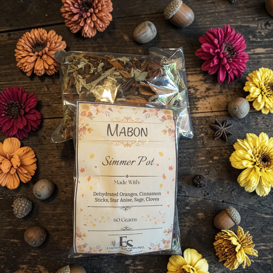 Mabon simmer pot herbs and spices on a rustic wooden surface, with a printed front label reading 'MABON Simmer Pot.' The scene is styled with orange and magenta flowers, yellow flowers, acorns, and star anise around the package.
