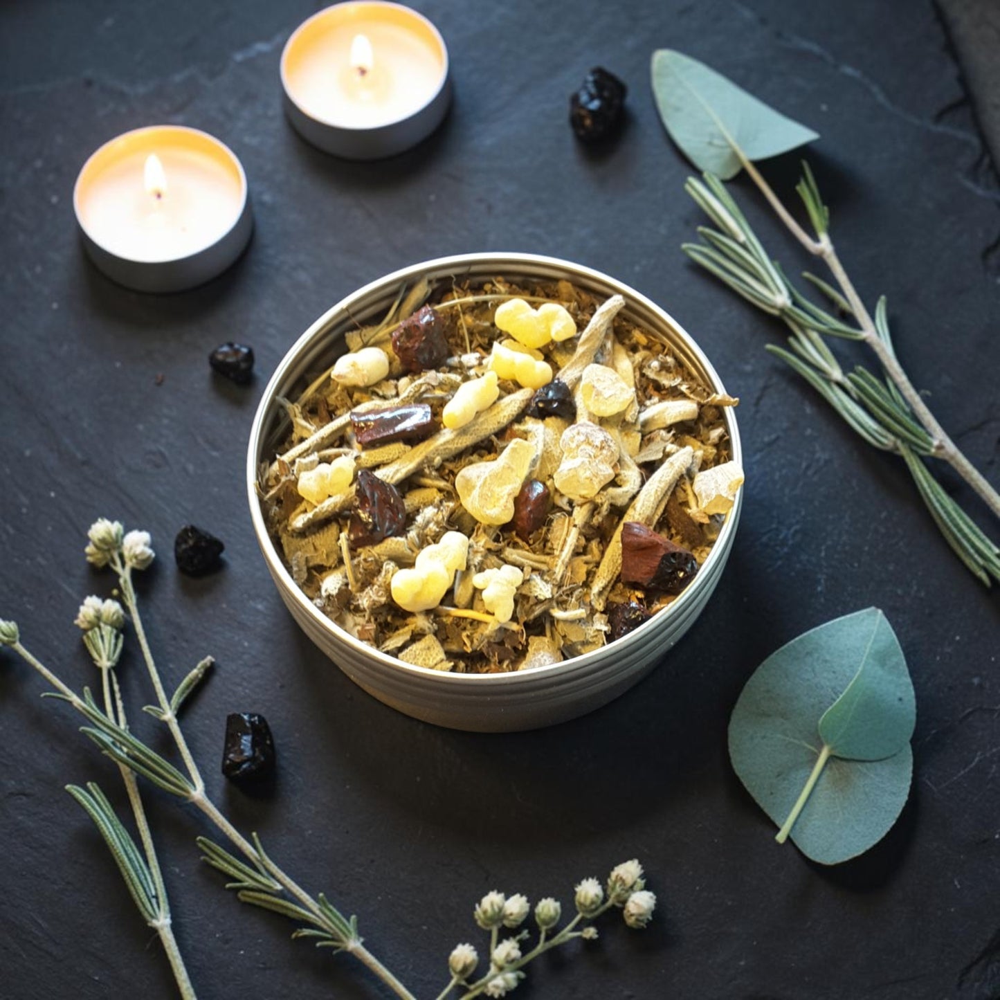 A blend of Samhain loose incense in a round metal tin, styled on a dark slate background with lit tea light candles, rosemary sprigs, eucalyptus leaves, and small black berries.