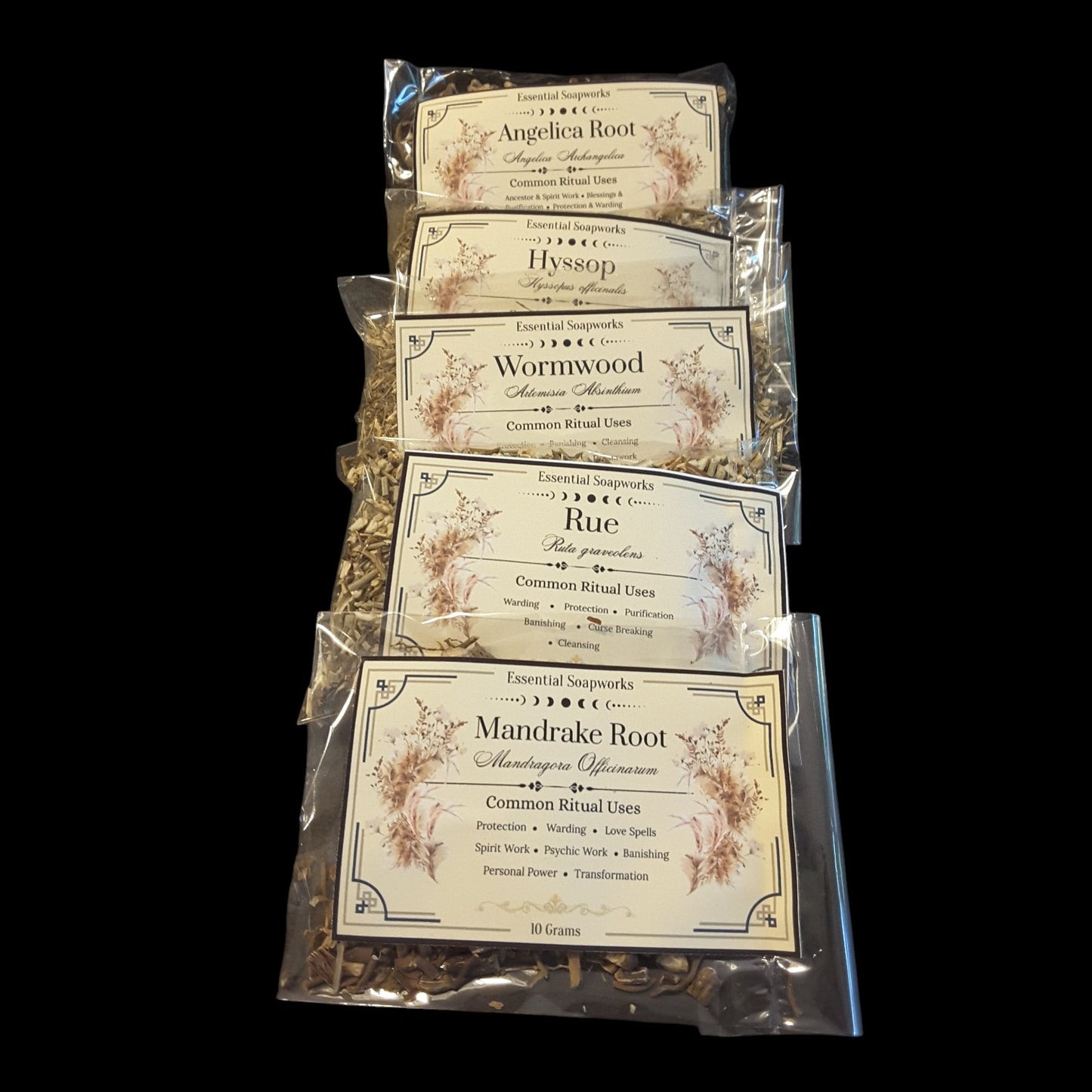 Five clear plastic bags of witchy Herbs for Banishing spells and rituals arranged in a vertical stack on a black background. Each bag has a cream-colored label with ornate floral borders and text including Angelica Root, Hyssop, Wormwood, Rue, and Mandrake Root.